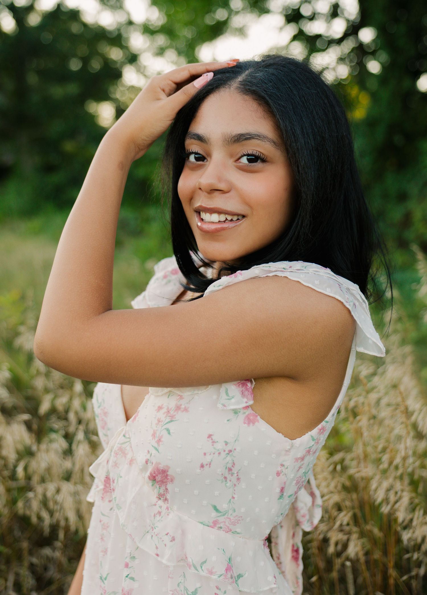 Young woman in floral dress smiling over shoulder in a sunlit field, hand in dark hair.