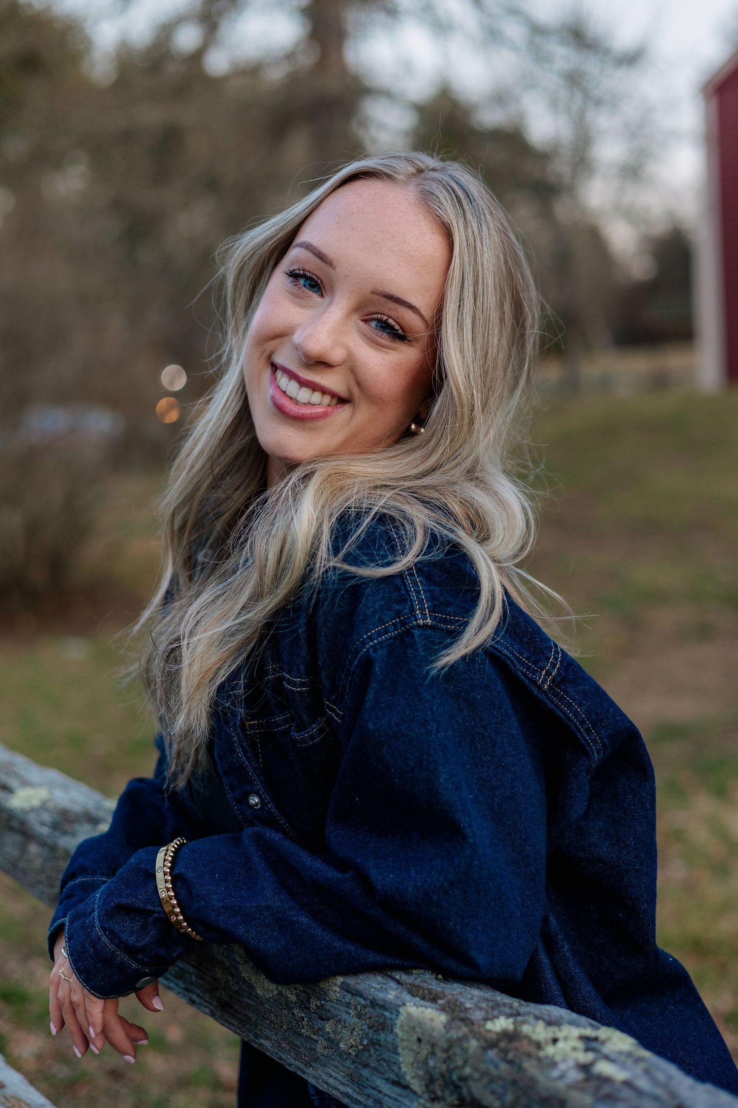 Smiling blonde woman in dark denim jacket leaning on weathered wooden fence in outdoor setting.