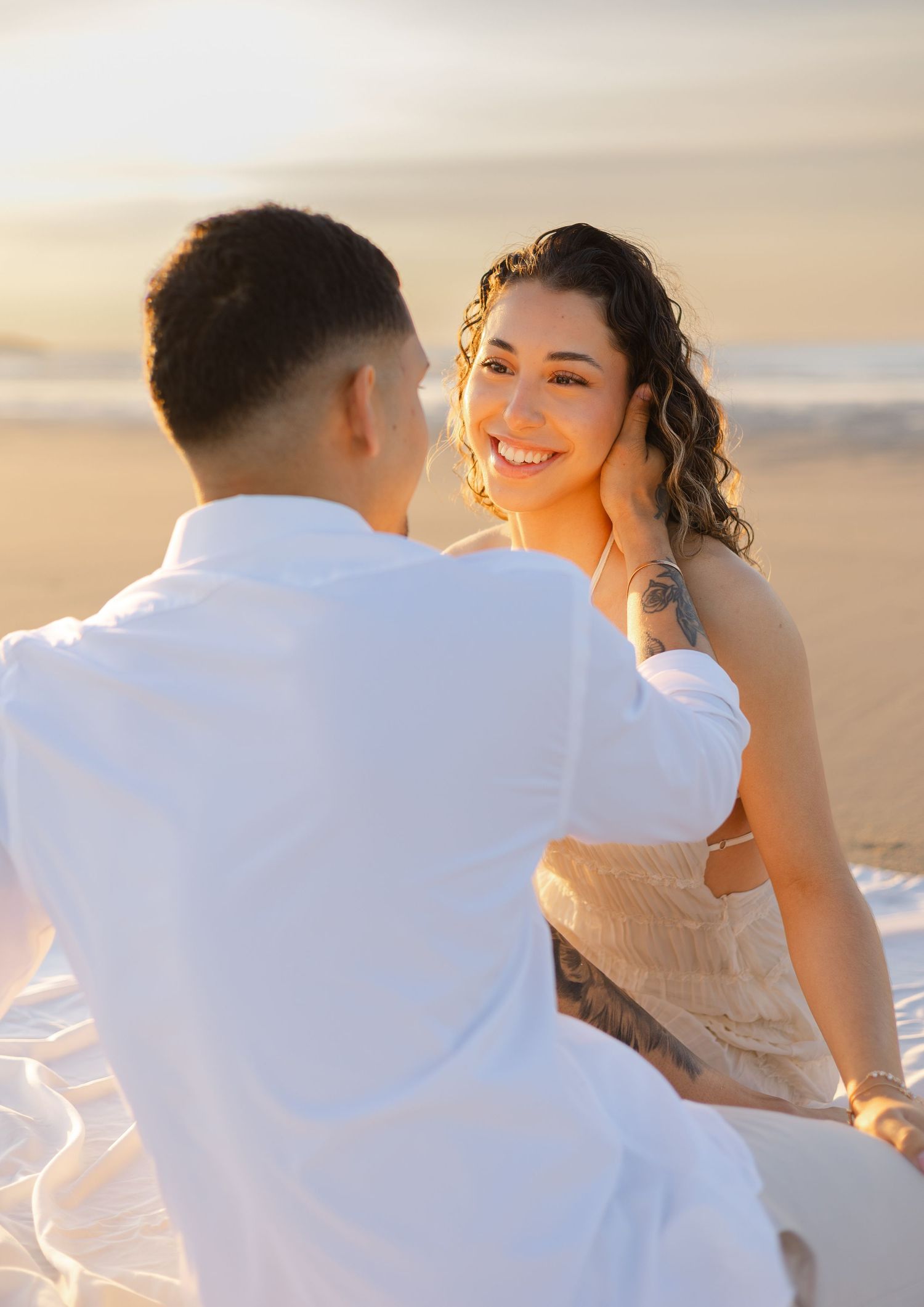 A smiling woman with curly hair and a tattoo sits on a beach at sunset with a man in a white shirt.