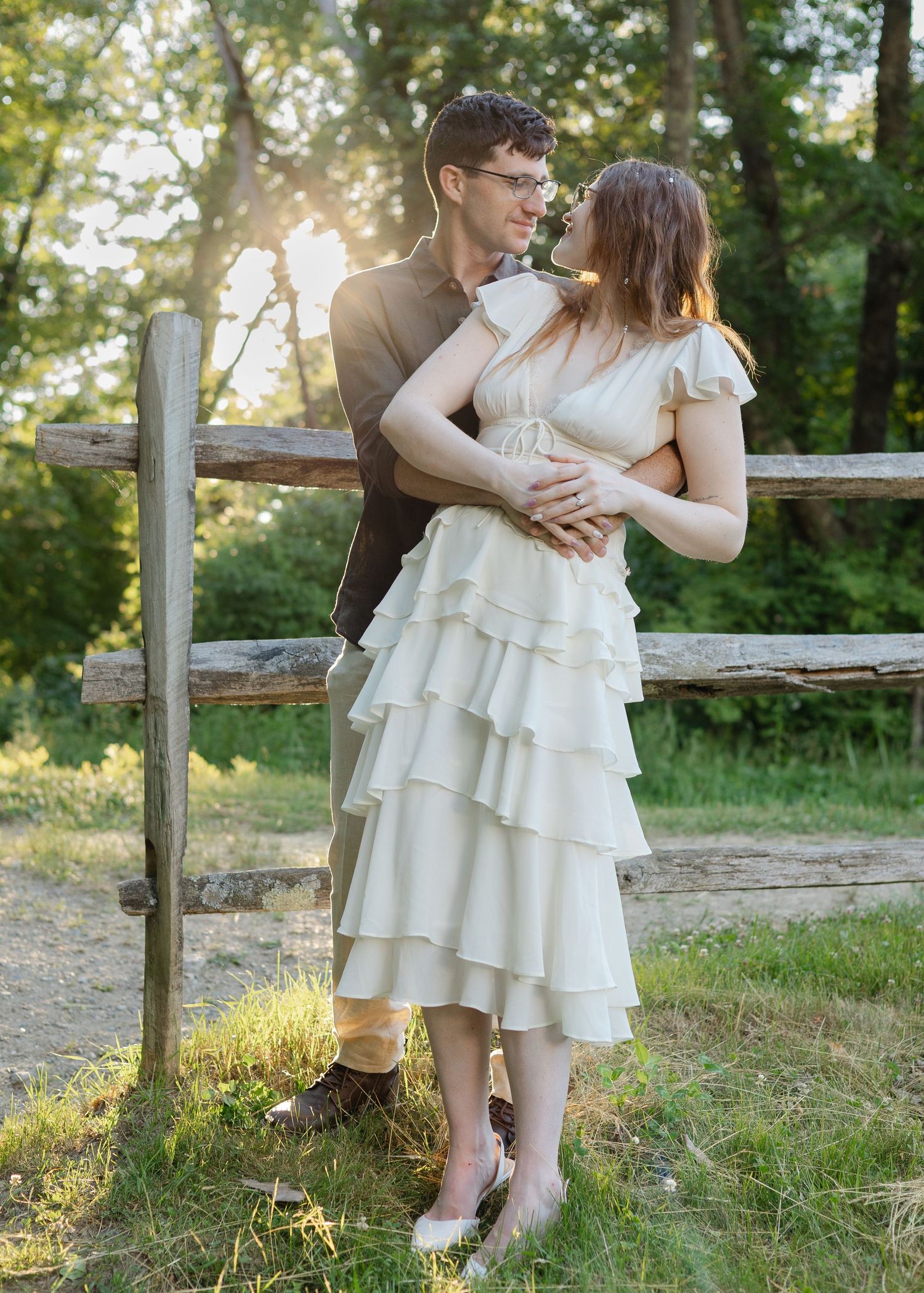 Couple embracing near rustic wooden fence in sunlit forest, woman in cream ruffled dress, man in brown suit.