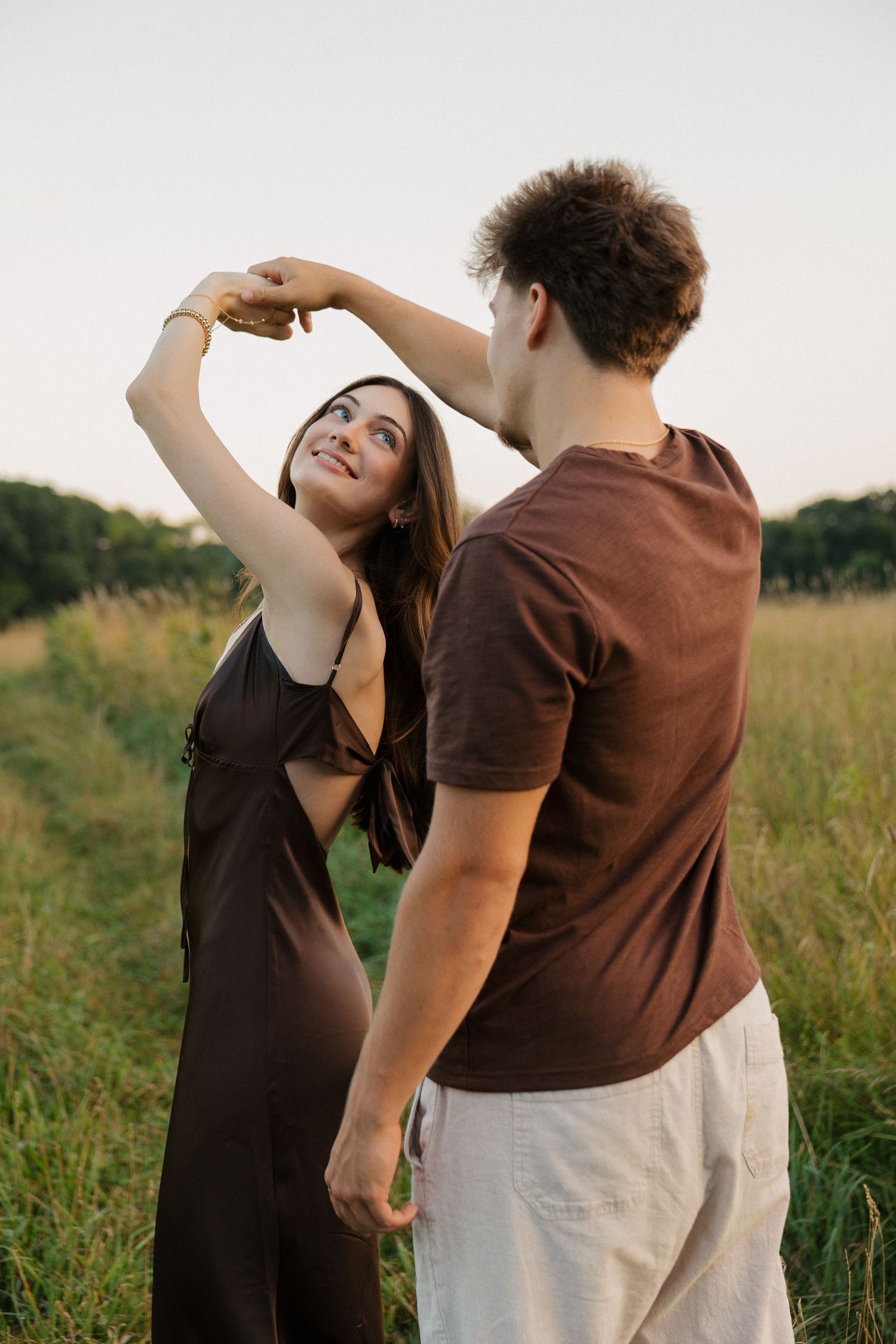 A couple dancing in a green field at sunset, woman in black dress smiling as man spins her around.