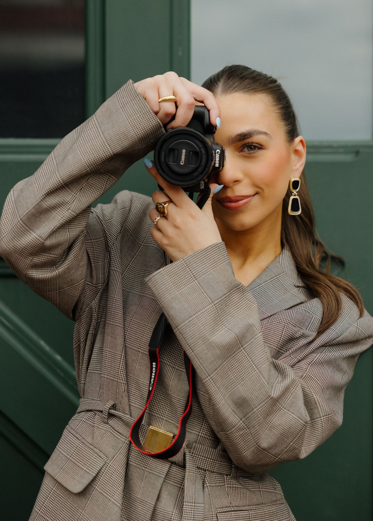Woman in plaid blazer holding a Canon camera up to her face, wearing gold rings and earrings, posed outdoors.