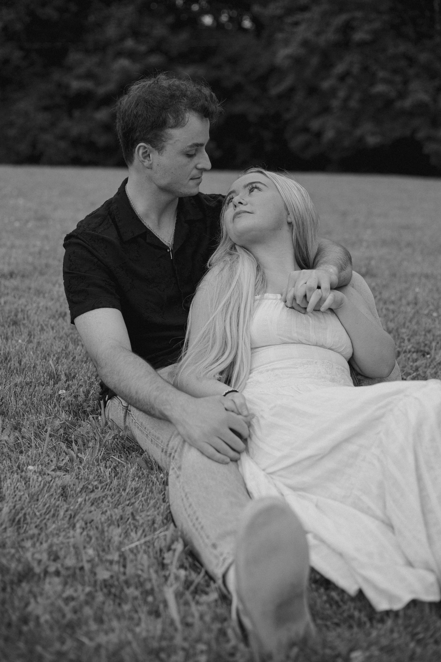 Black and white photo of a couple sitting on grass, woman in white dress leaning against man in black shirt.