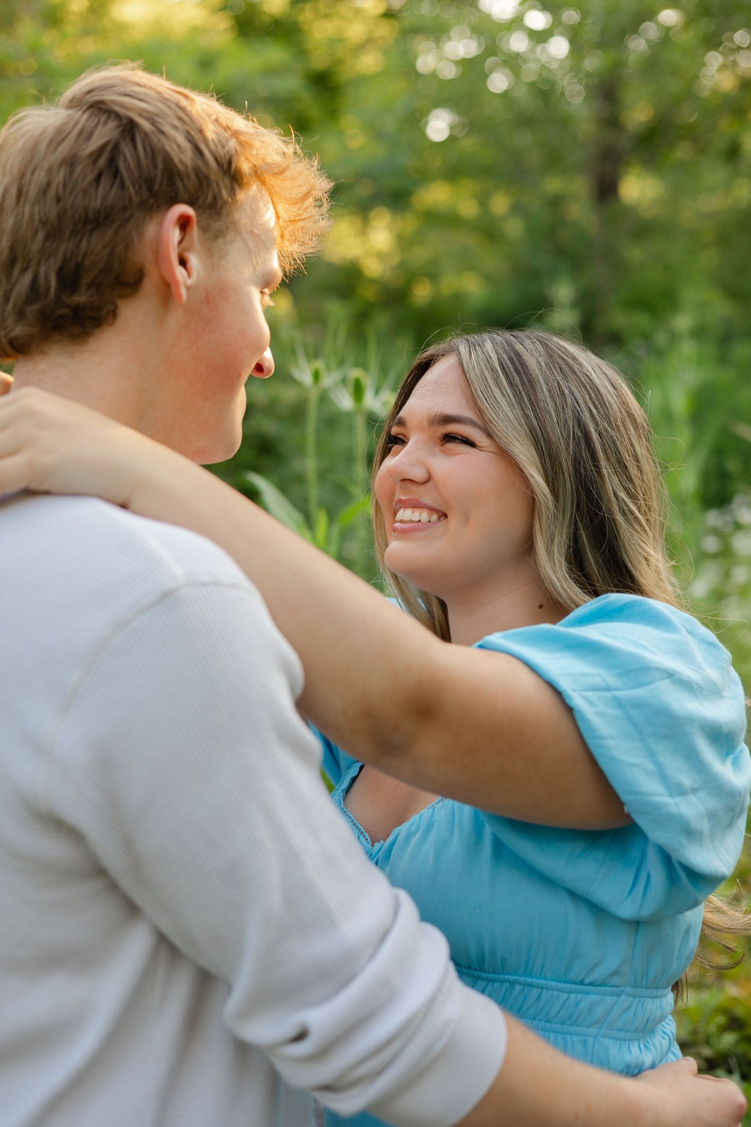 A smiling woman in a blue dress embraces a man outdoors, surrounded by lush green foliage.