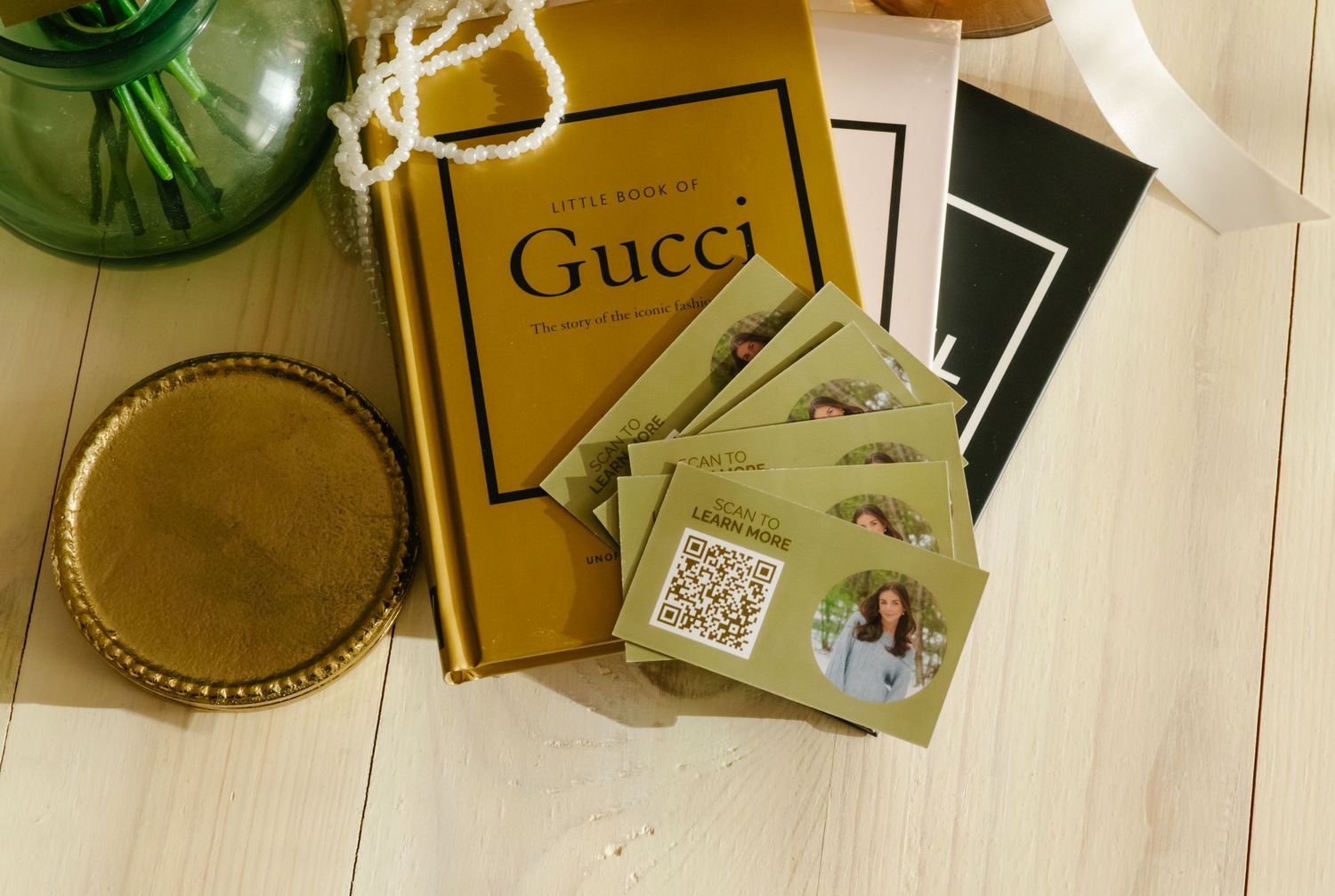 Gucci coffee table book with QR code business cards, gold tray, and green vase on white wood surface.