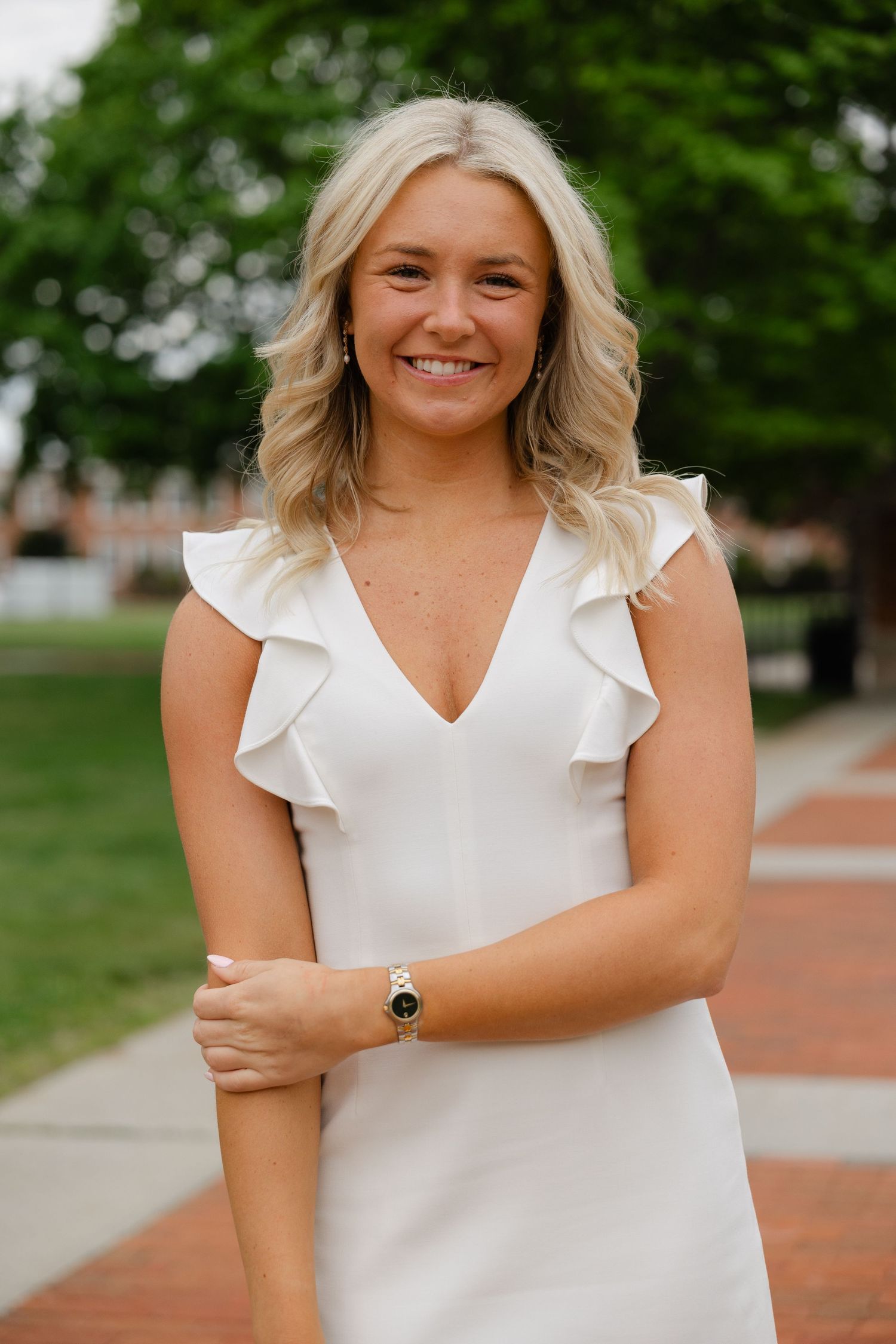 Smiling young woman in white ruffle dress with gold watch, posing outdoors on brick pathway with green trees.