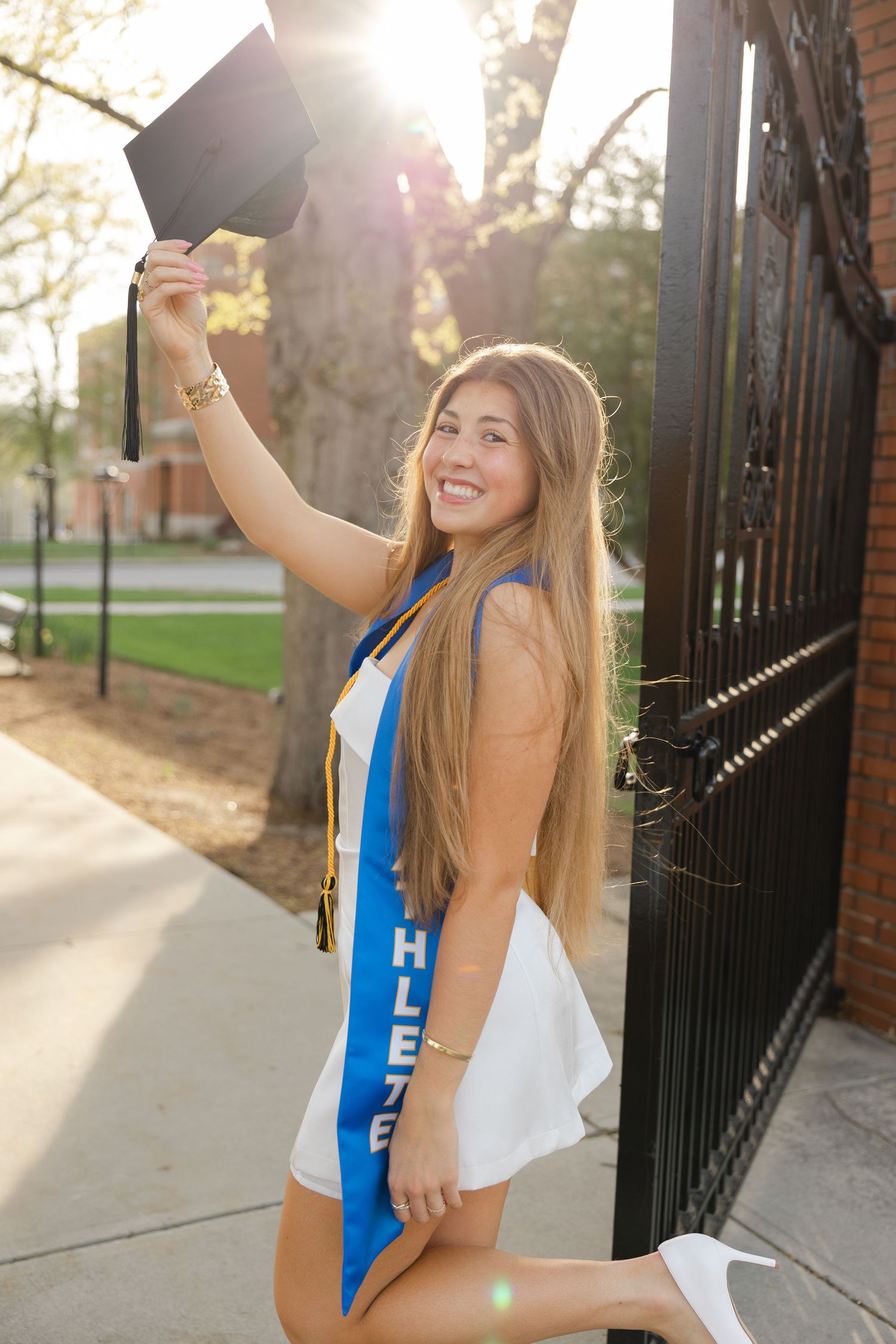 Smiling graduate in white dress holds cap aloft, wearing blue athlete sash, posing by ornate iron gate on sunny campus.