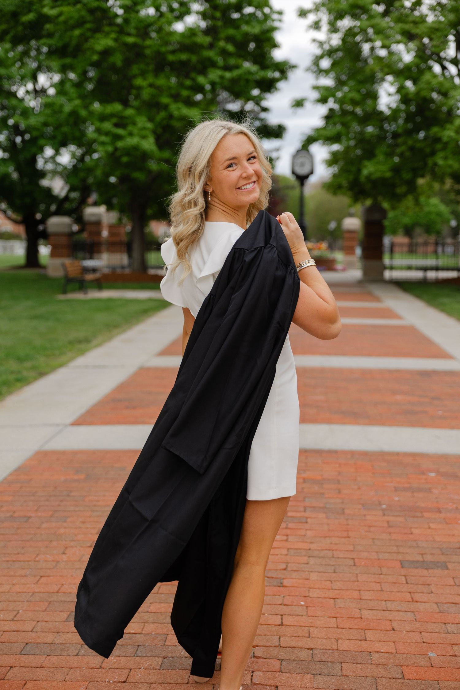 Smiling blonde graduate in white dress holds black graduation gown over shoulder on brick campus walkway.