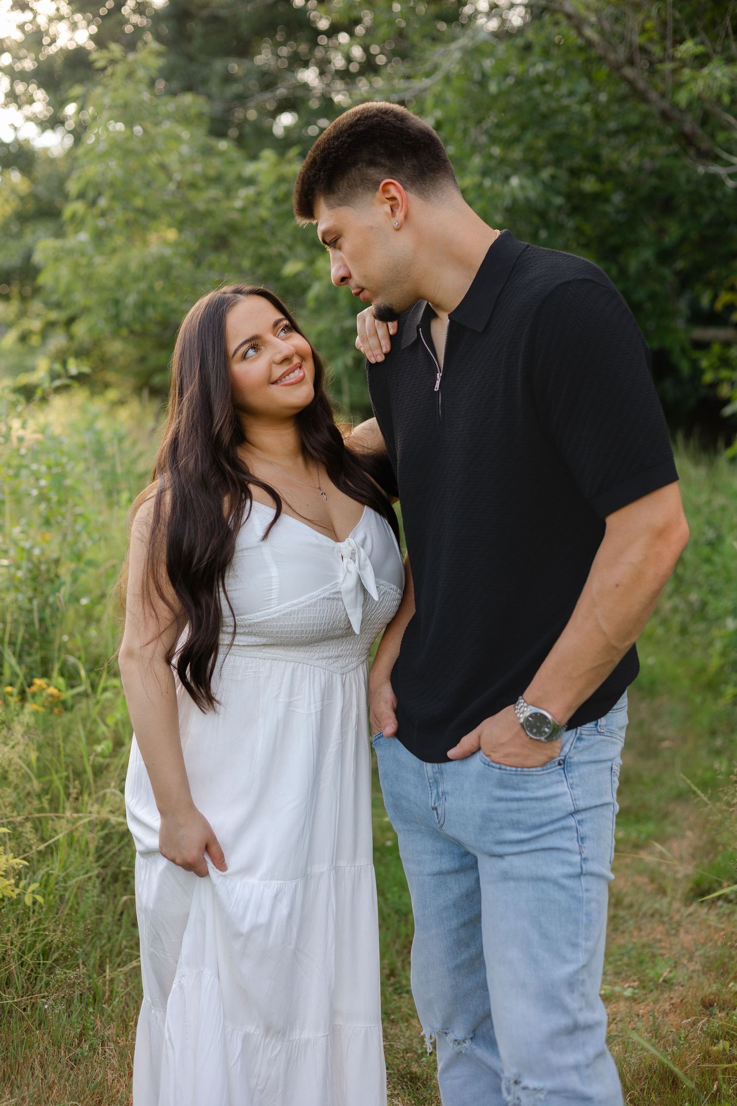 A couple poses outdoors, woman in white dress smiling up at man in black shirt and jeans, surrounded by lush greenery.