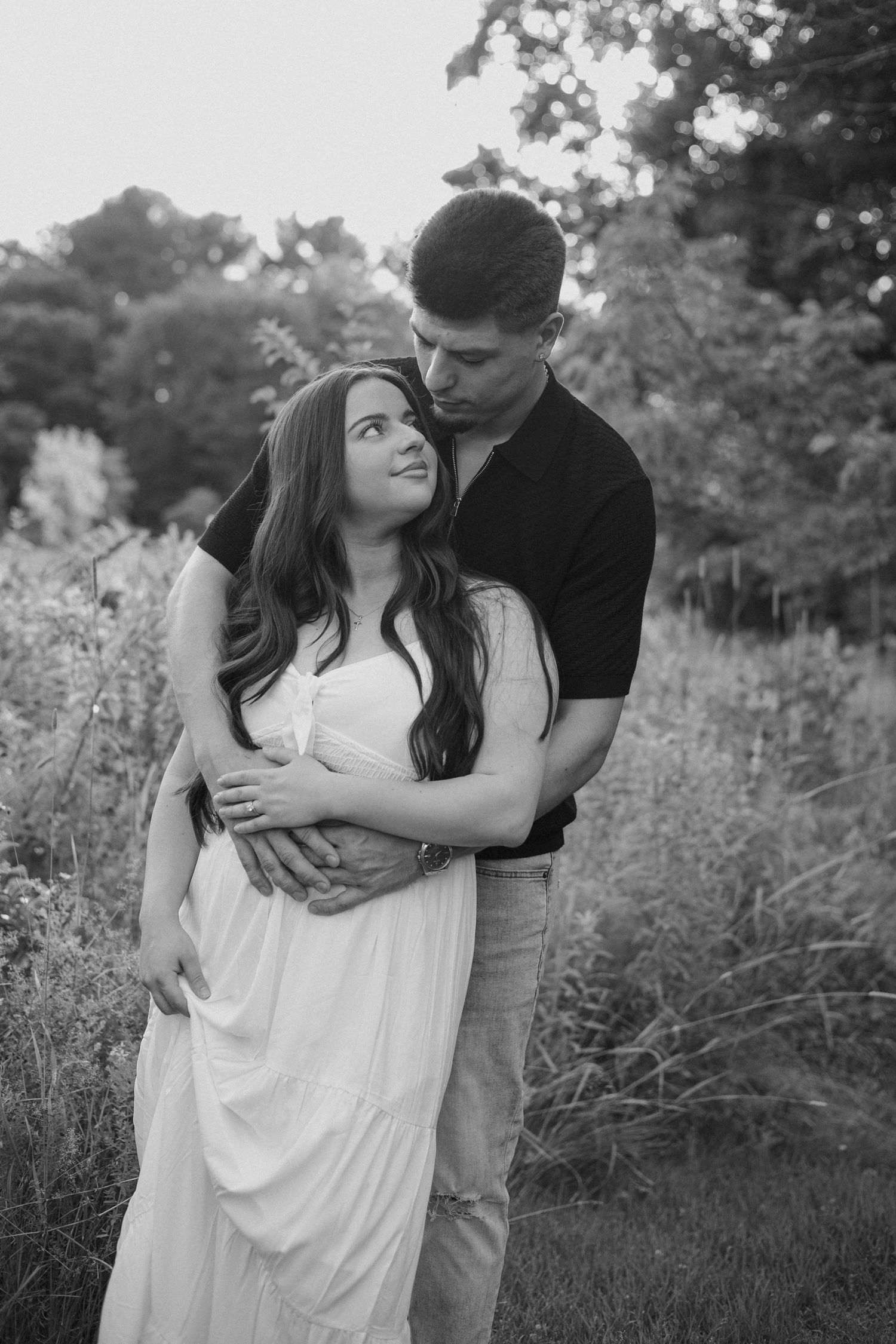 Black and white photo of a couple embracing outdoors, woman in white dress looking up at man surrounded by lush greenery.