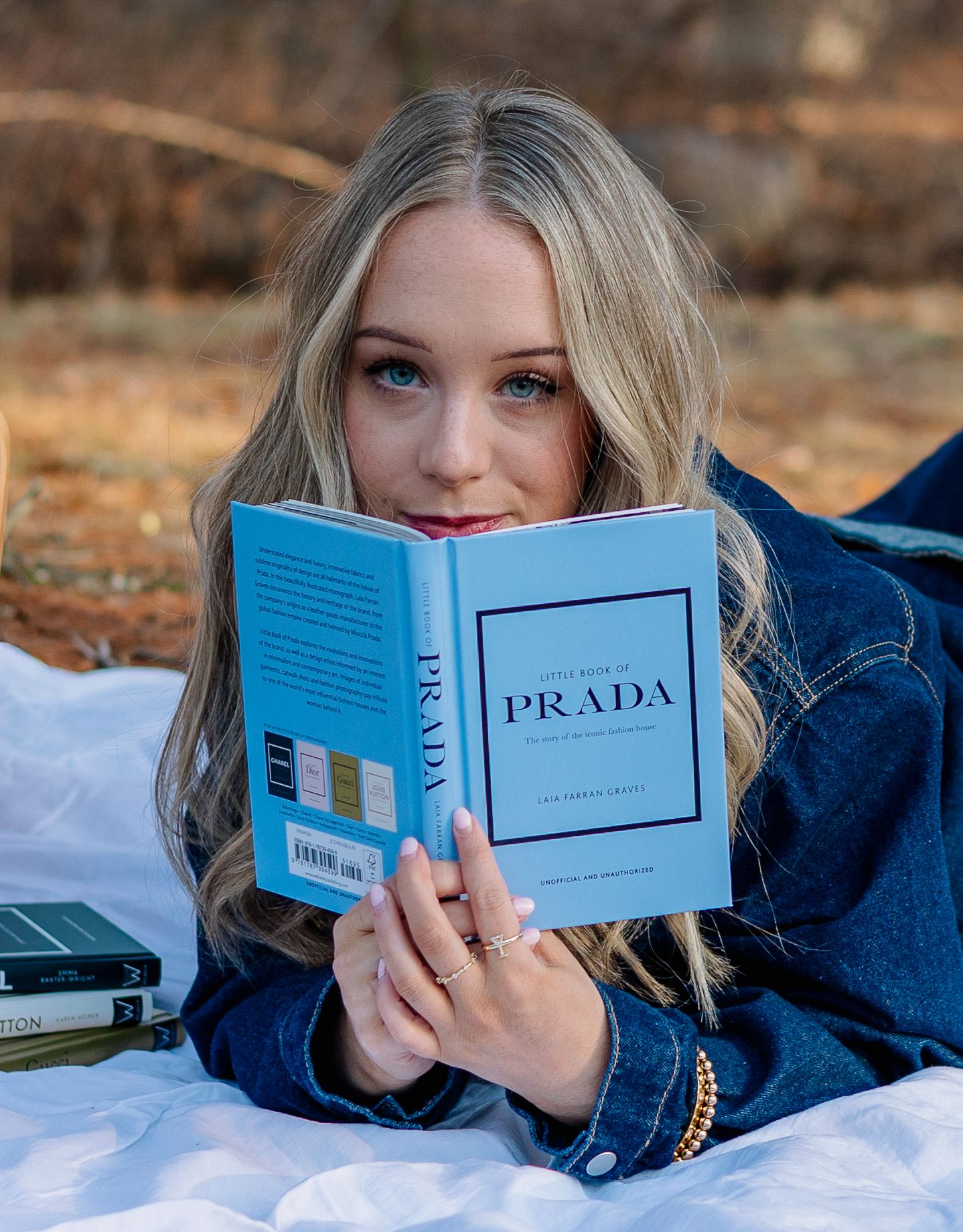 Young blonde woman reading a little book of Prada outdoors on a white blanket in a denim jacket.