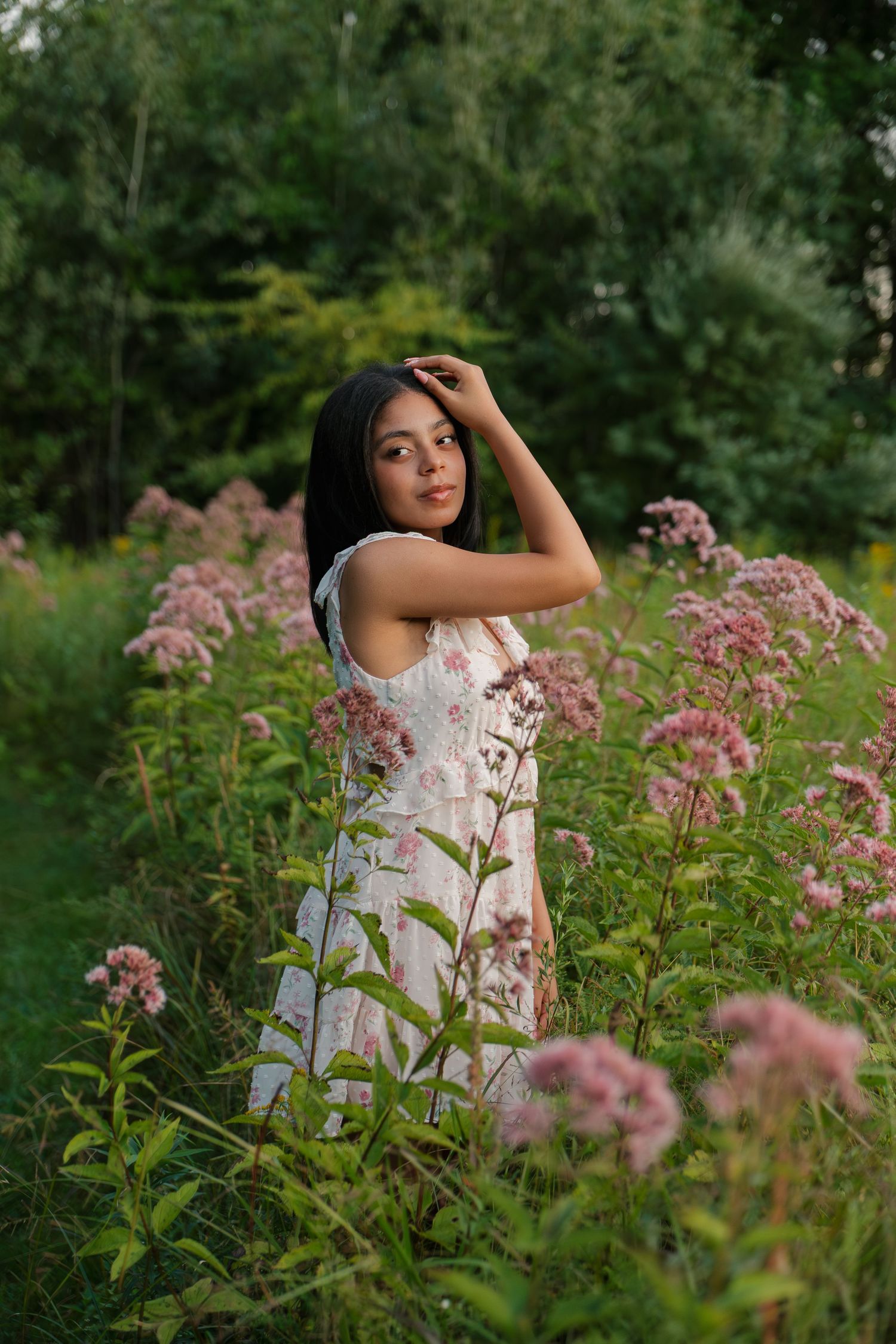Young woman in floral dress posing among pink wildflowers in a lush green garden setting.