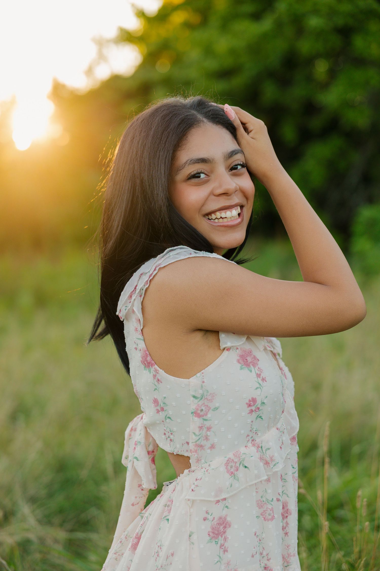 Smiling young woman in floral dress poses outdoors at golden hour with lush green background.