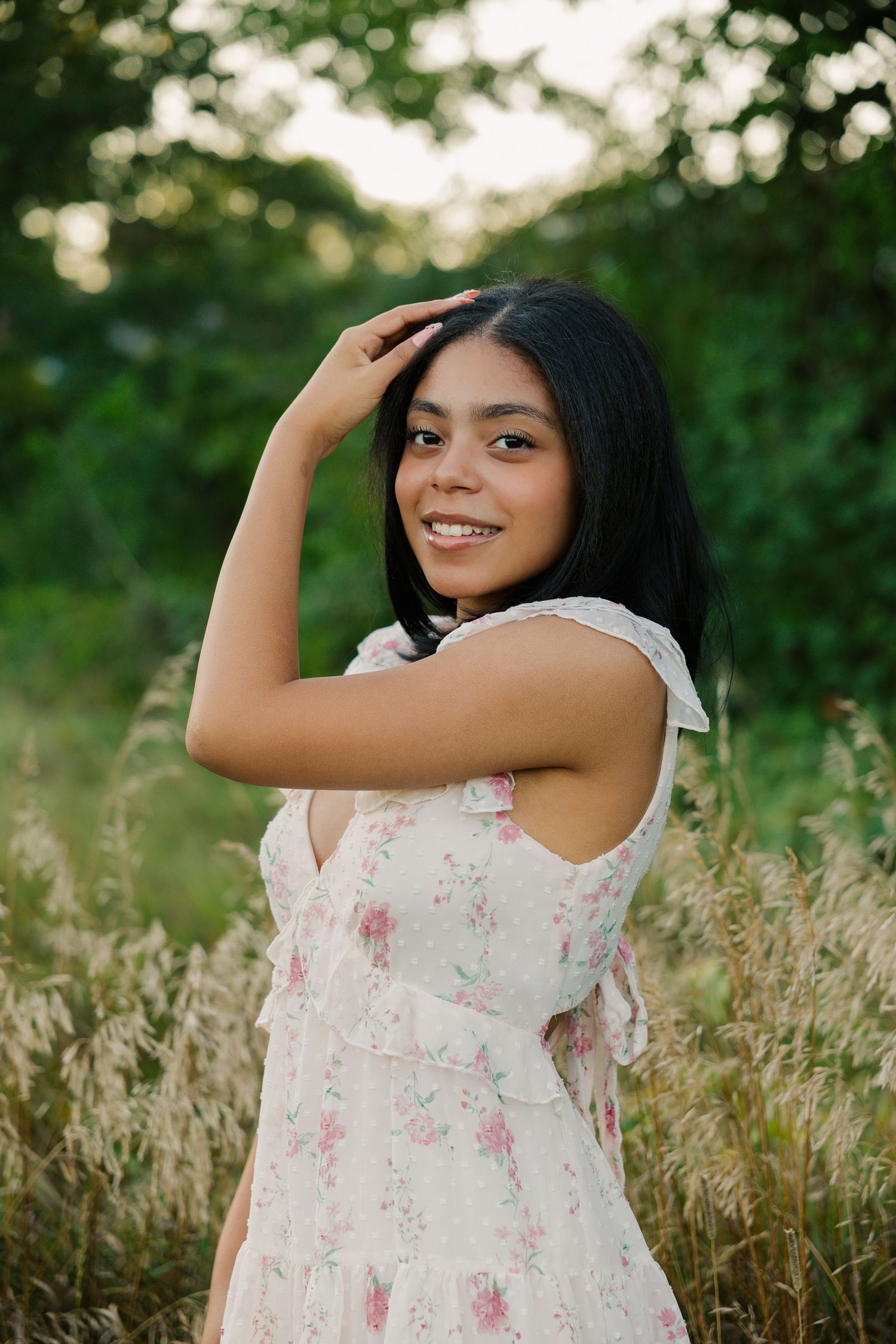 Young woman in floral dress smiling outdoors in tall grass with green trees in background.