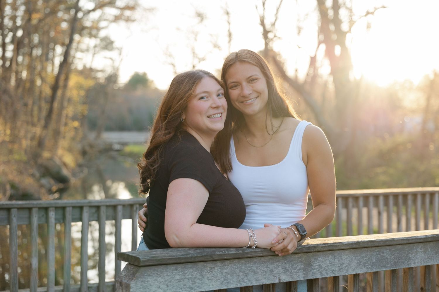Two smiling young women leaning on a wooden bridge railing at sunset.