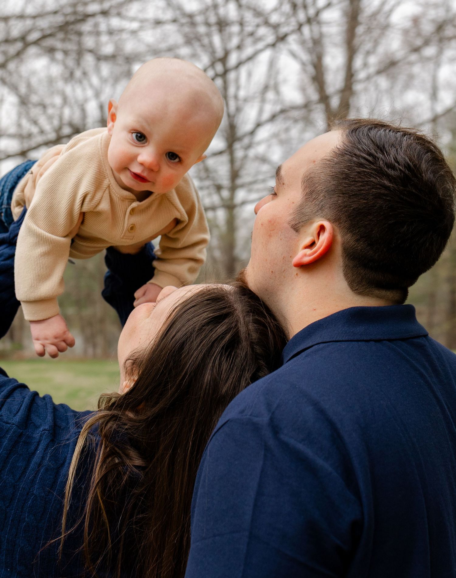 Parents lovingly holding baby up in the air outdoors among bare winter trees.