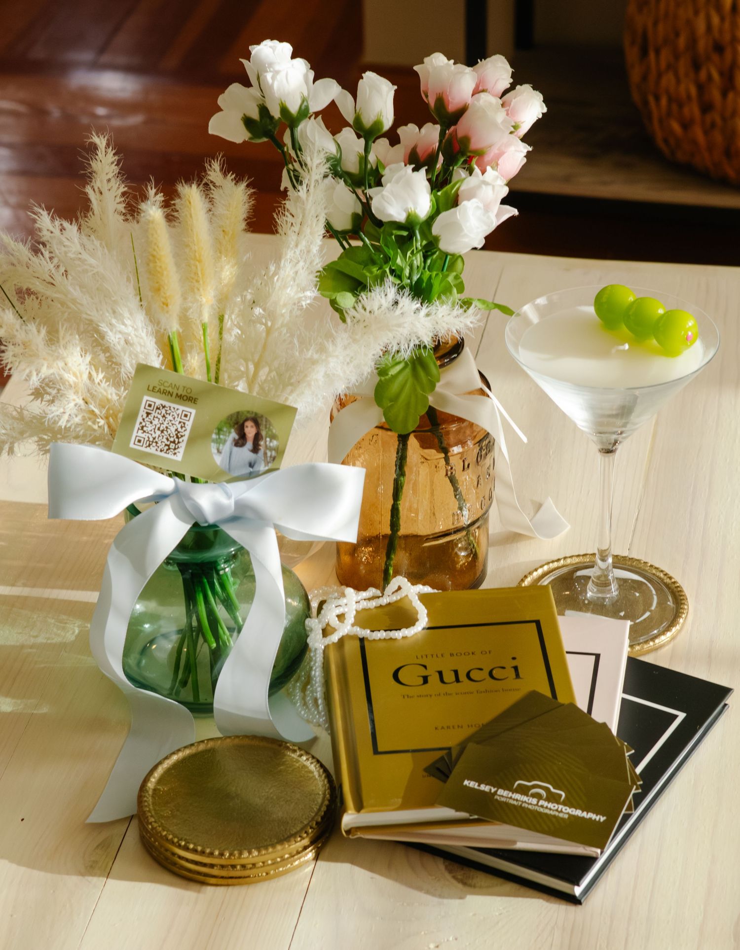 Elegant table display with white roses, Gucci books, a martini glass, gold coasters, and a ribbon-tied green vase arrangement.