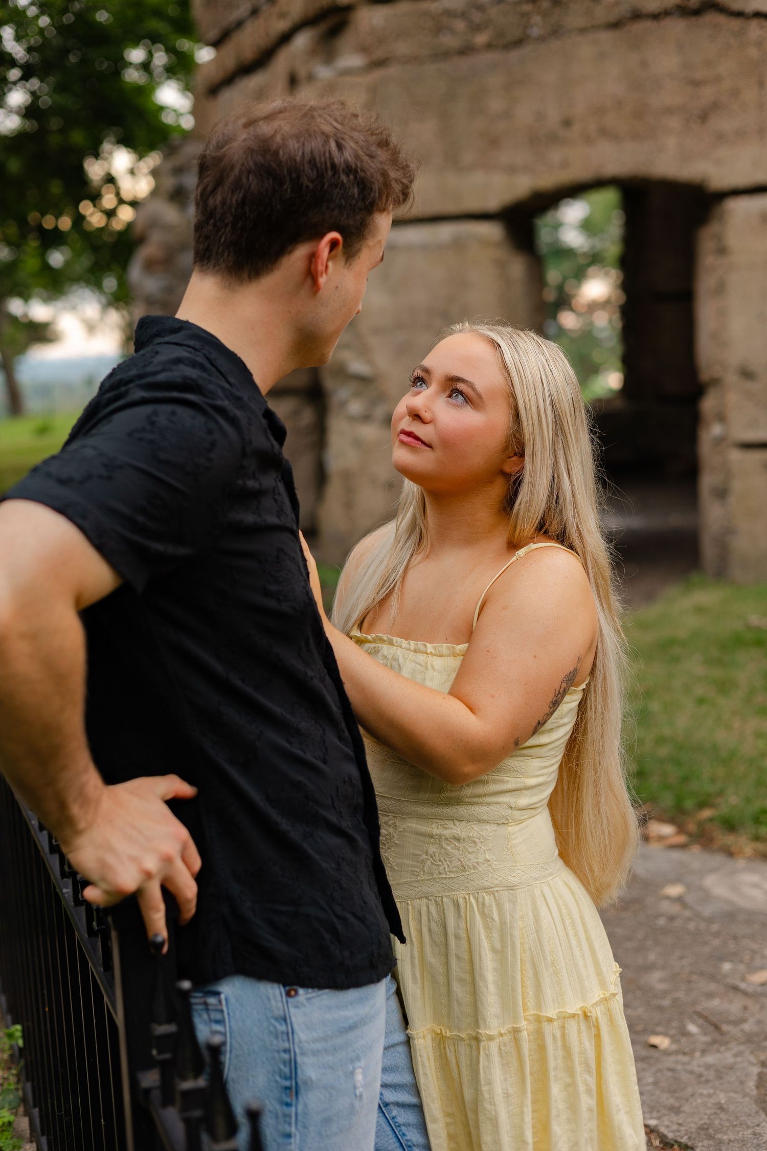 A young couple shares a romantic moment outdoors, with the blonde woman in a yellow dress gazing up at her partner.