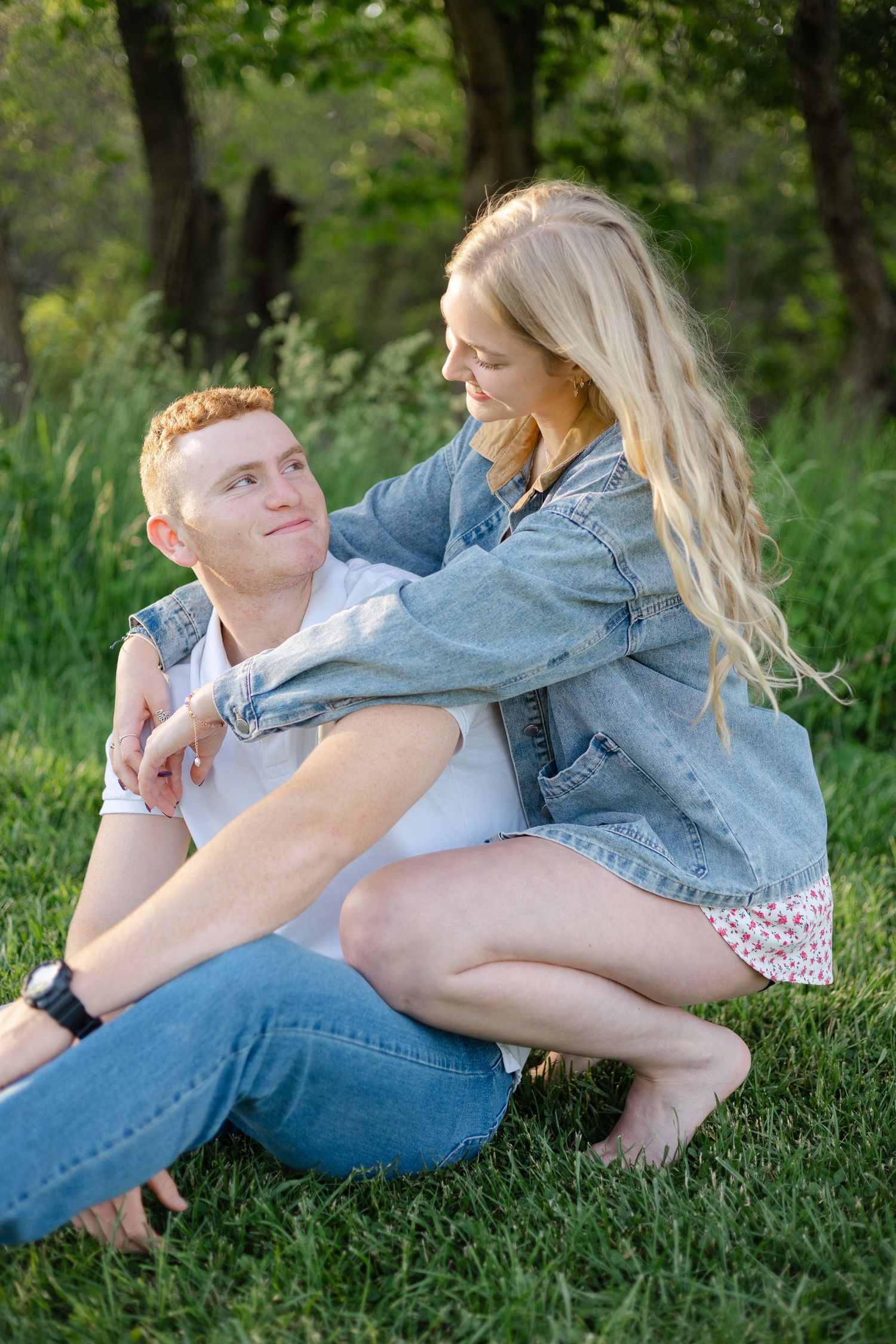 Couple sitting on grass in park, woman in denim jacket leaning over smiling man at golden hour.