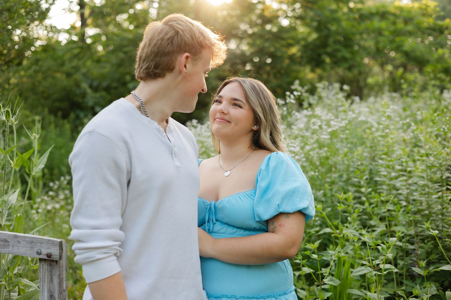 A young couple smiles at each other in a sunlit green field with wildflowers during a romantic outdoor photo session.