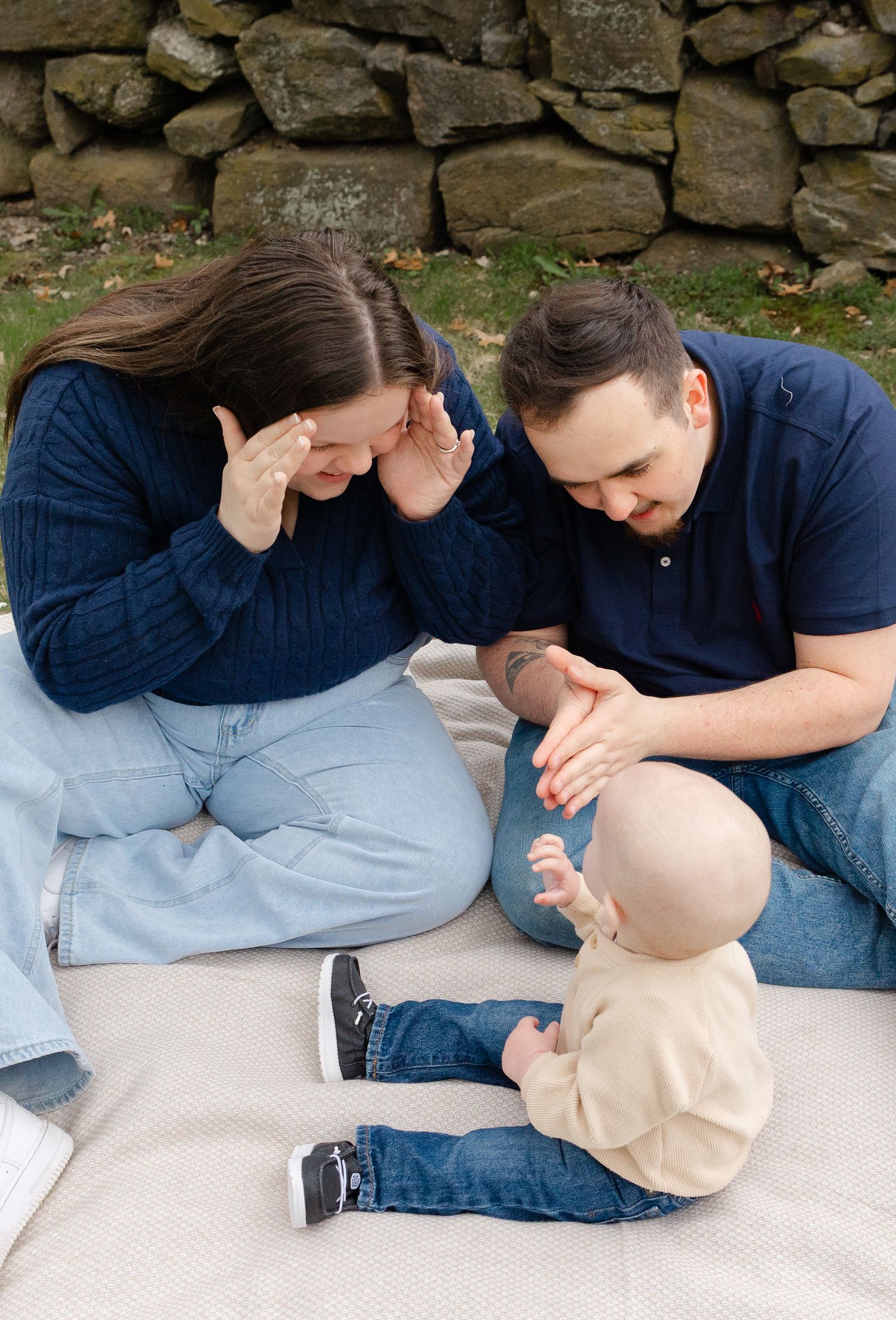 A young family sits on a blanket outdoors, parents smiling at their baby boy near a stone wall.