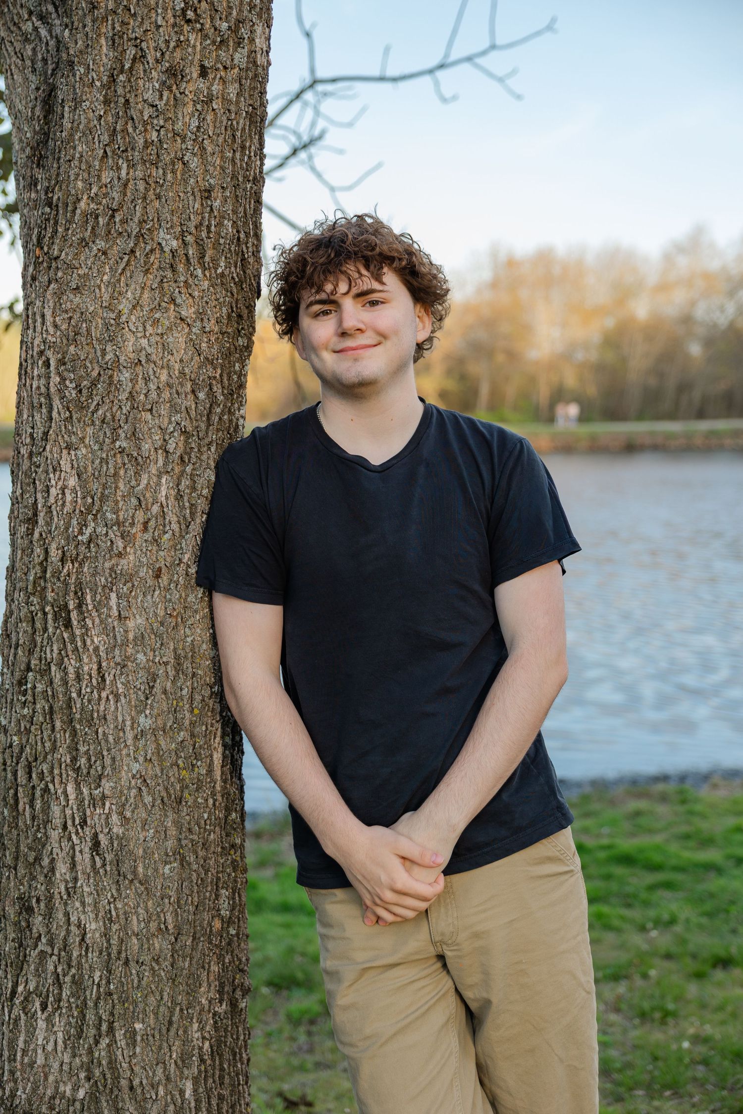 Young man with curly hair in black t-shirt leaning against tree by lake, smiling outdoors.