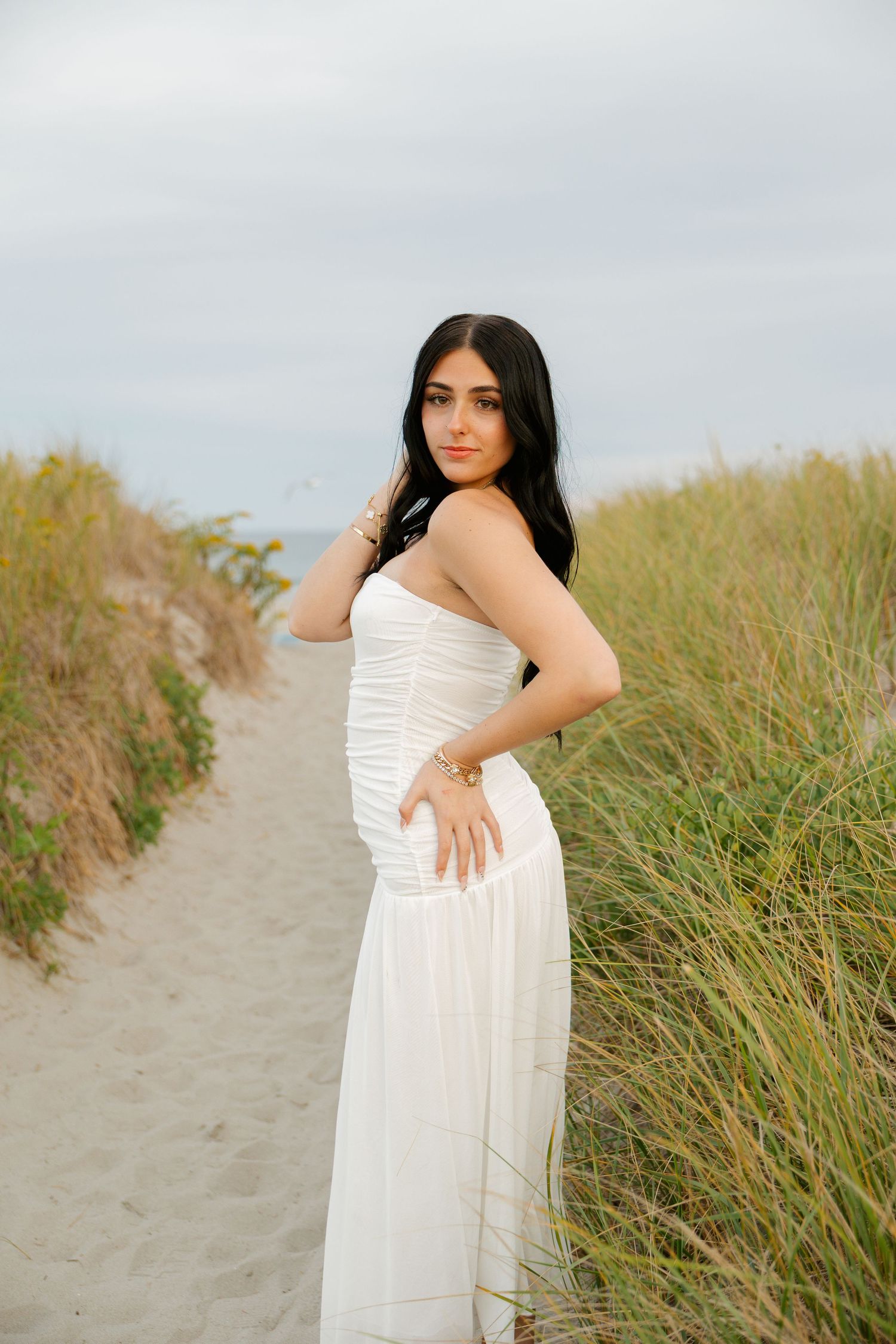 Young woman in white strapless dress poses on sandy beach path surrounded by tall grass under cloudy sky.