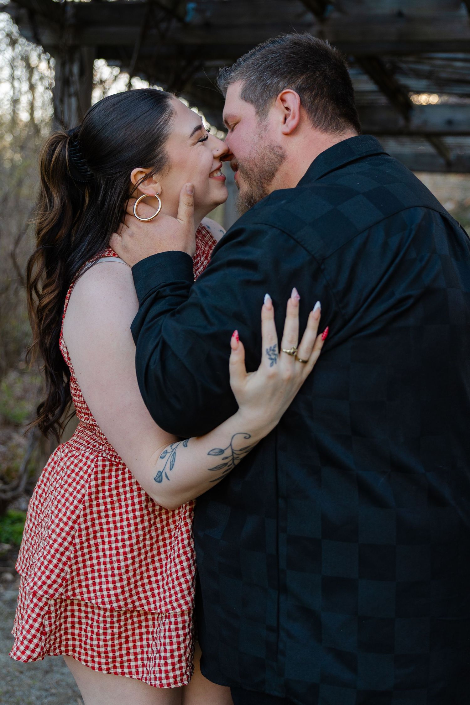 A couple shares a romantic kiss outdoors, the woman in a red gingham dress with tattoos and gold earrings.