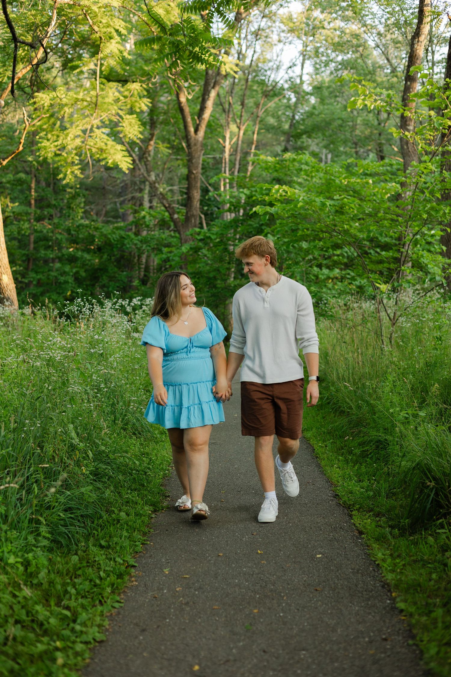 Couple holding hands walking on a lush green tree-lined path, woman in blue dress and man in casual summer outfit.