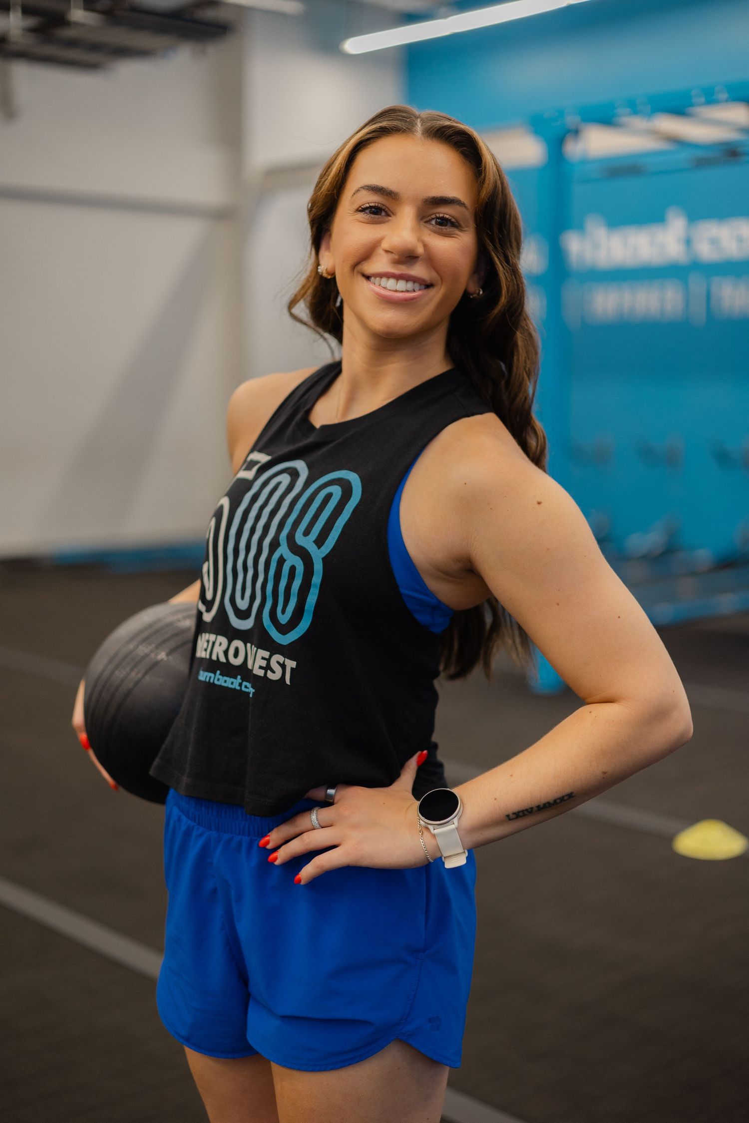 Woman in black tank top and blue shorts holds medicine ball and smiles in a modern gym setting.