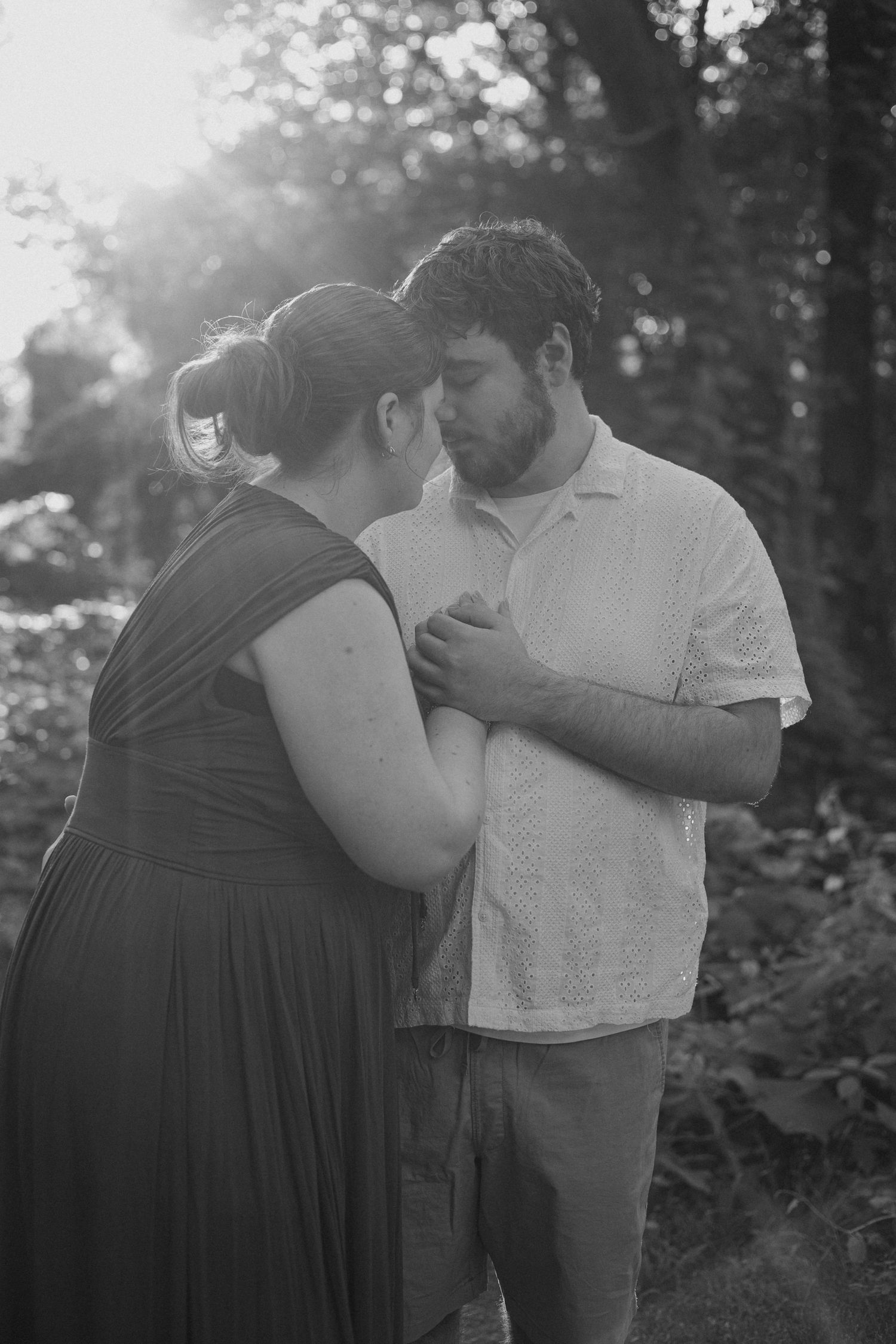 Black and white photo of a couple sharing an intimate moment outdoors, with soft backlit sunlight filtering through trees.