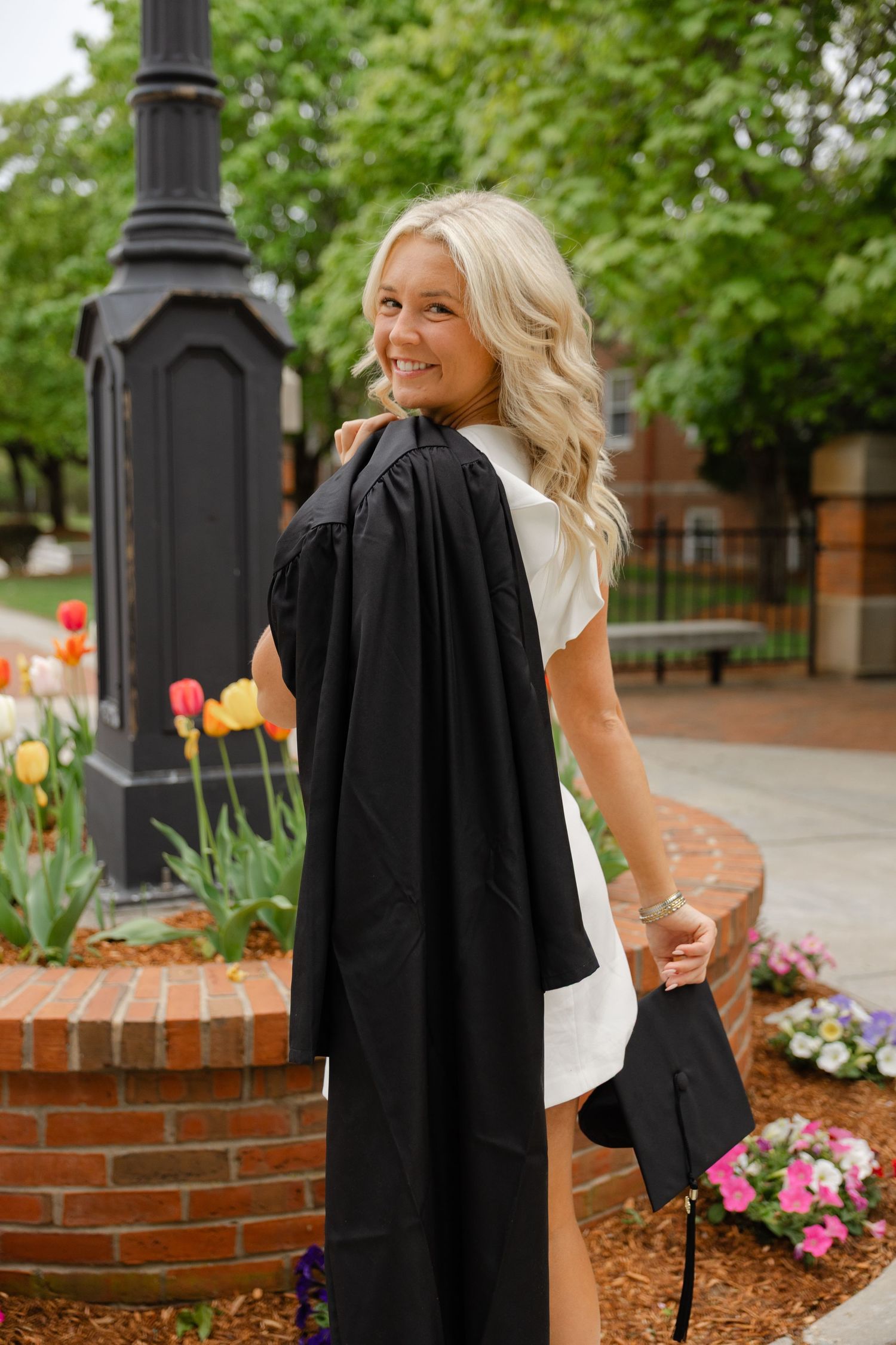 Blonde graduate smiling over shoulder wearing black gown and white dress near colorful tulips.