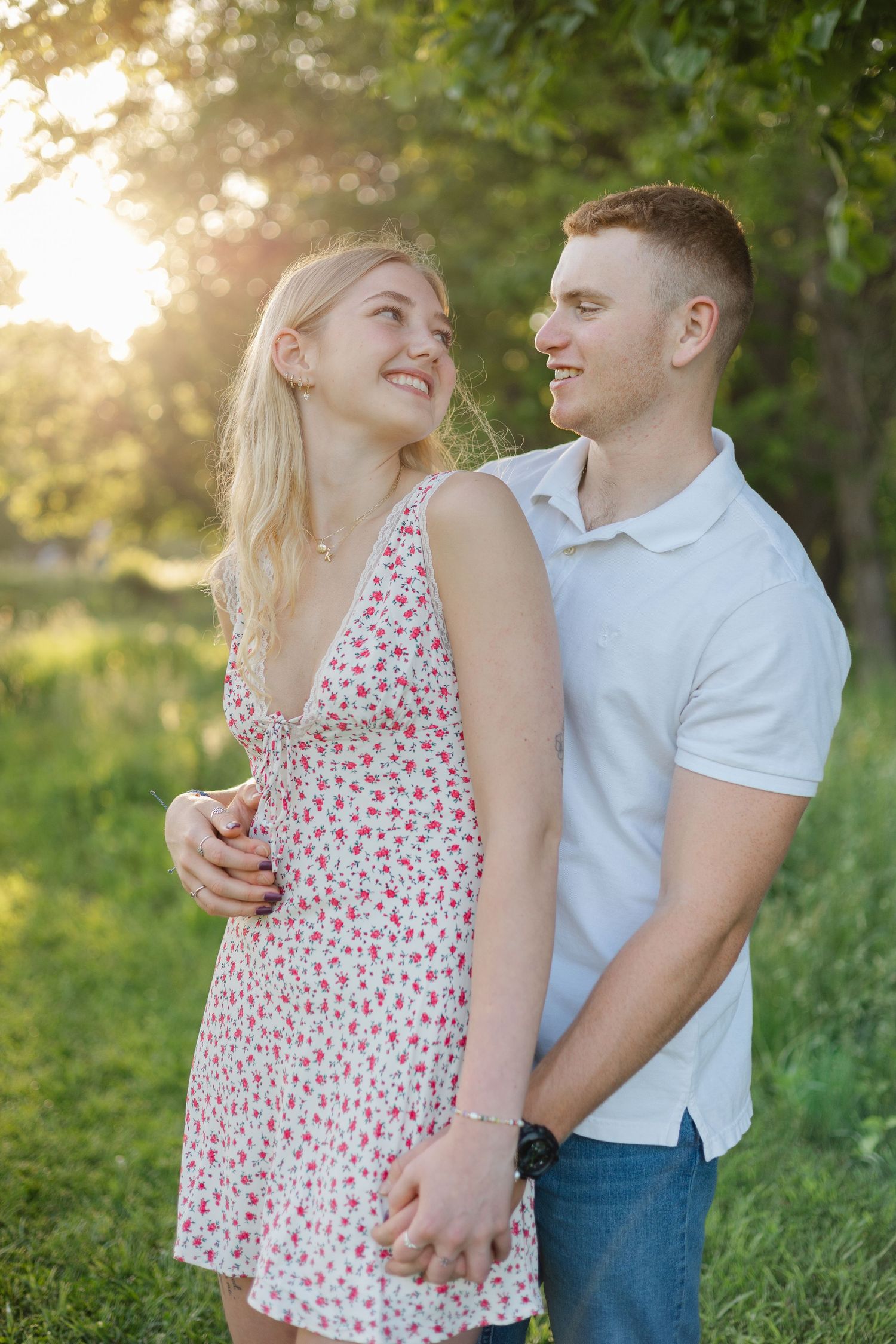 Smiling couple holding hands in golden sunset light, woman in floral sundress with man in white polo.