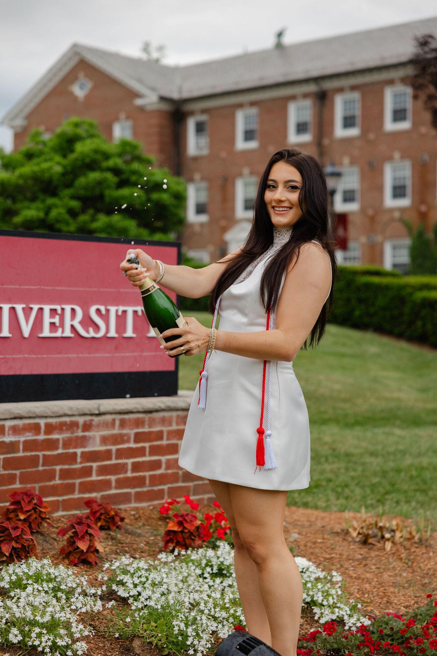 Celebrating female graduate popping champagne bottle in front of university sign in white dress.