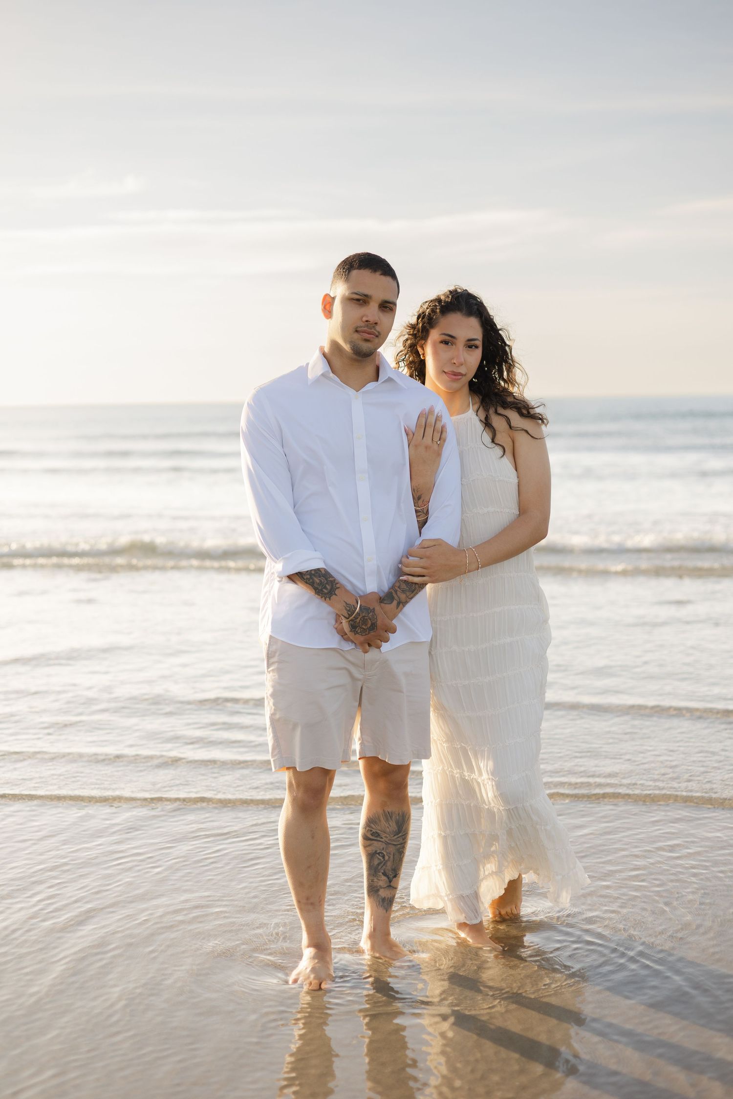 Couple in white outfits standing barefoot together on sandy beach at sunset holding hands.