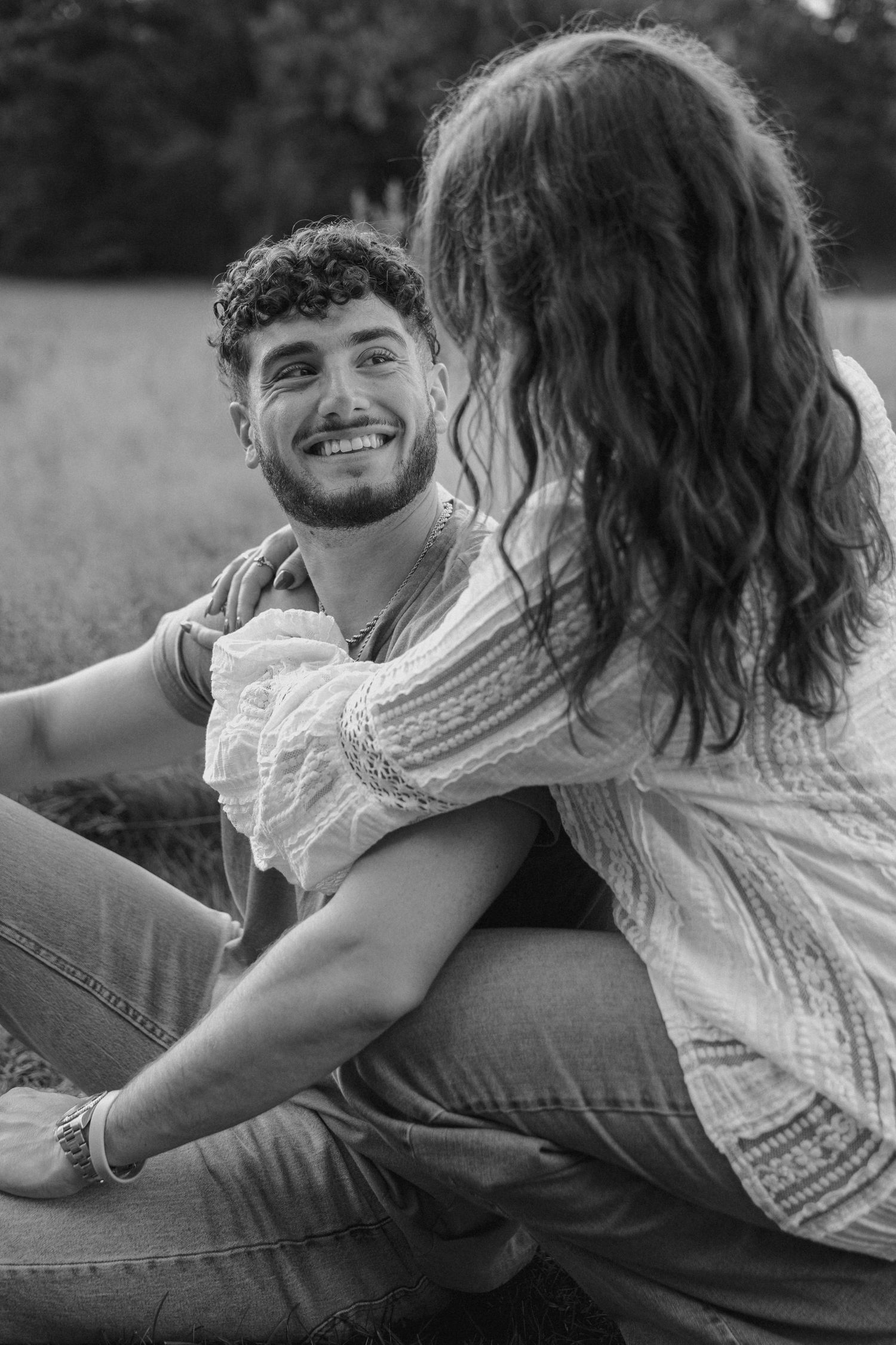 Black and white photo of a smiling man with curly hair sitting outdoors with a woman in a lace top.