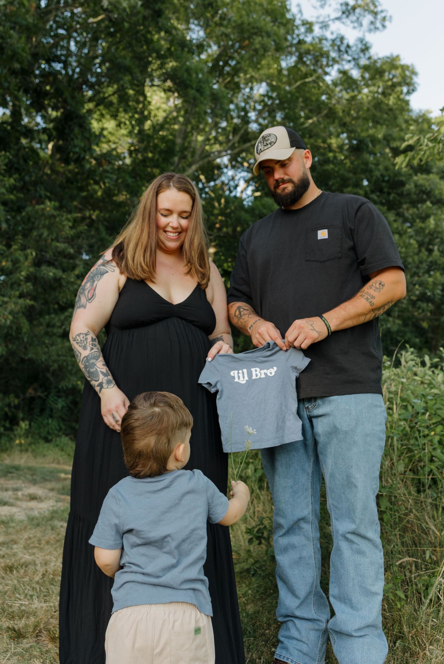 Pregnant woman in black dress with tattooed man holding lil bro onesie and toddler outdoors for gender reveal photo.
