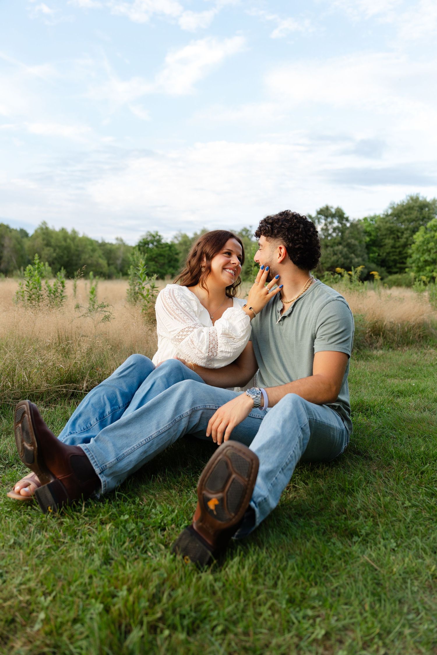 A couple sits on green grass in a field, smiling at each other, wearing jeans and casual tops on a cloudy day.