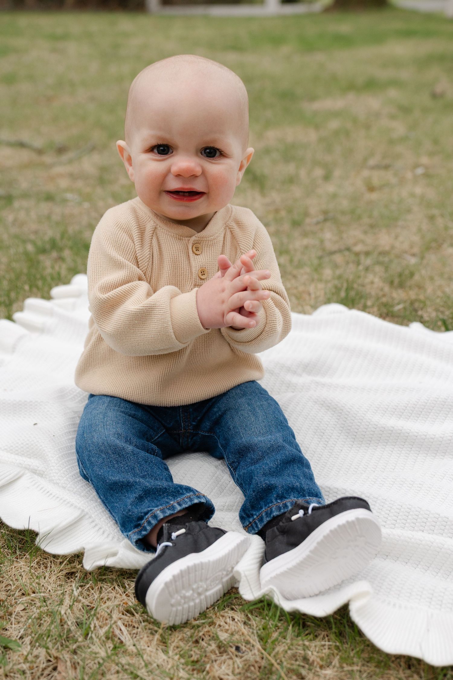 Adorable baby in beige sweater and jeans sitting on white blanket on grass, smiling.