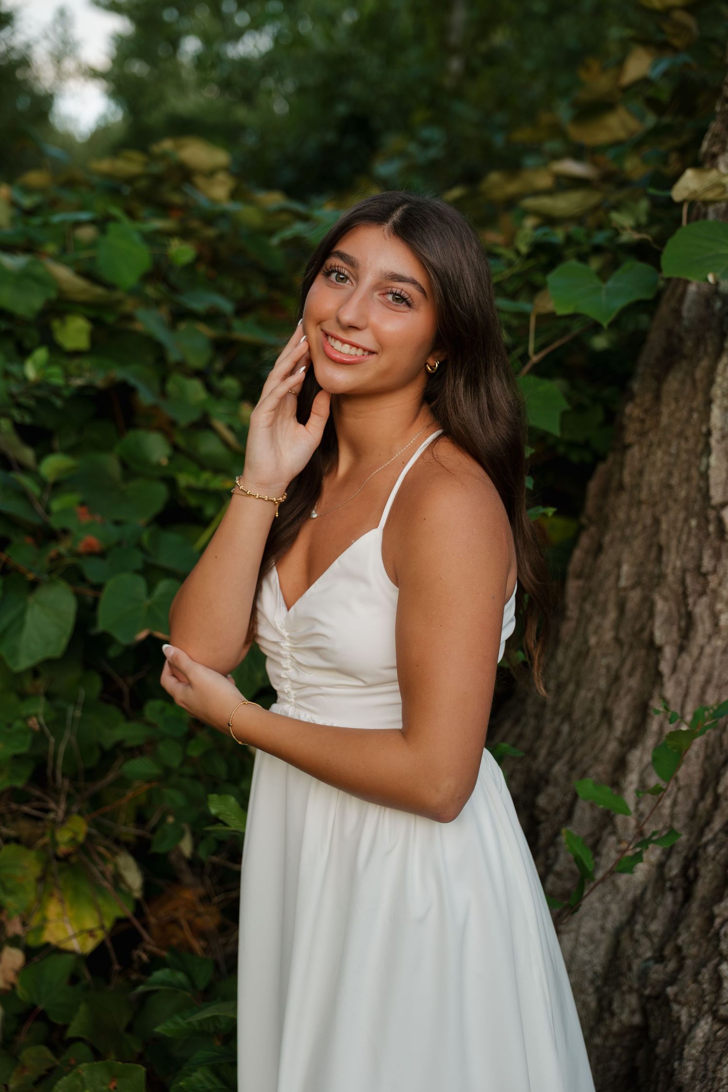 Young woman in white sundress smiling outdoors near tree and green foliage.