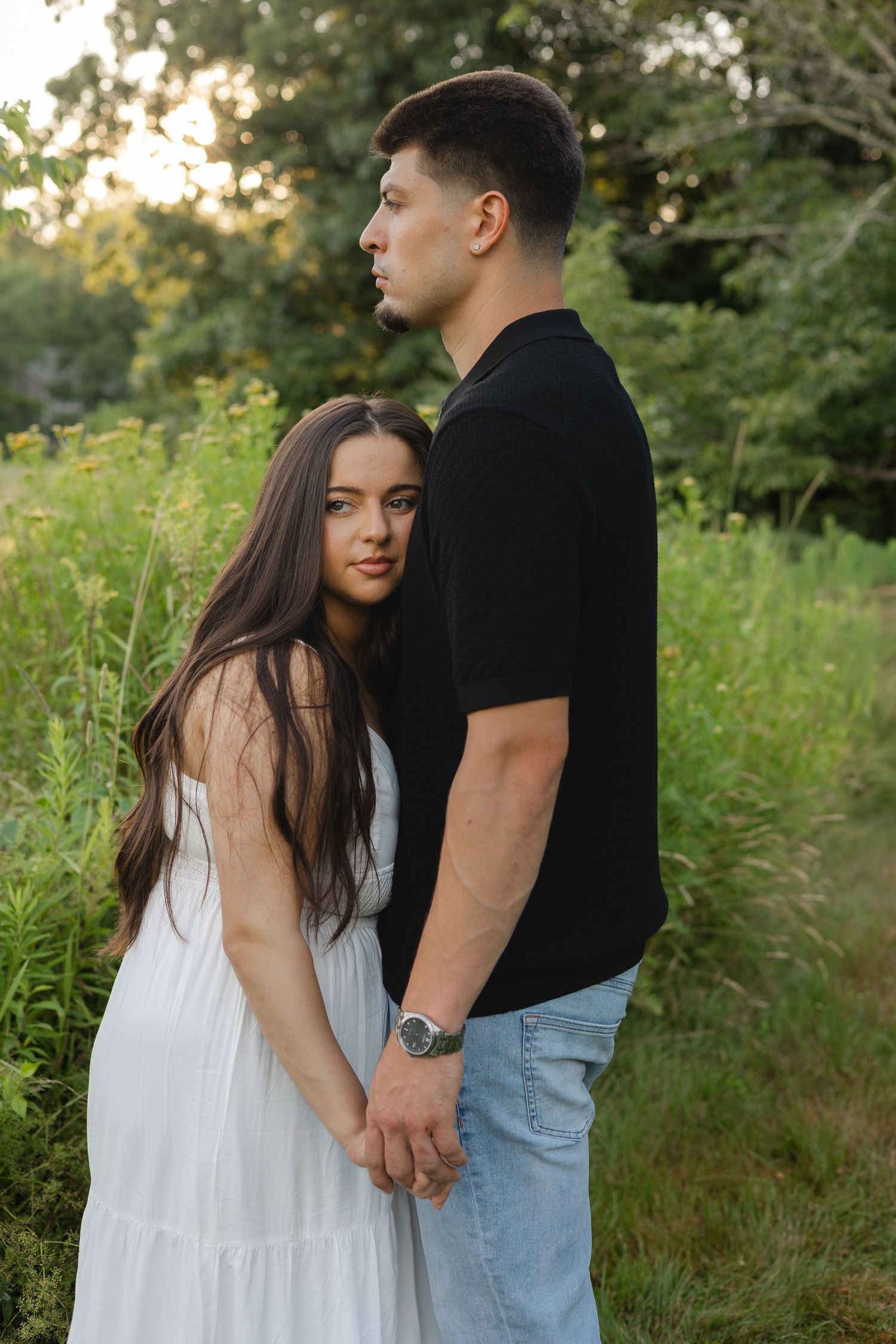 A couple holds hands outdoors, woman in white dress with long hair, man in black shirt and jeans, green nature background.