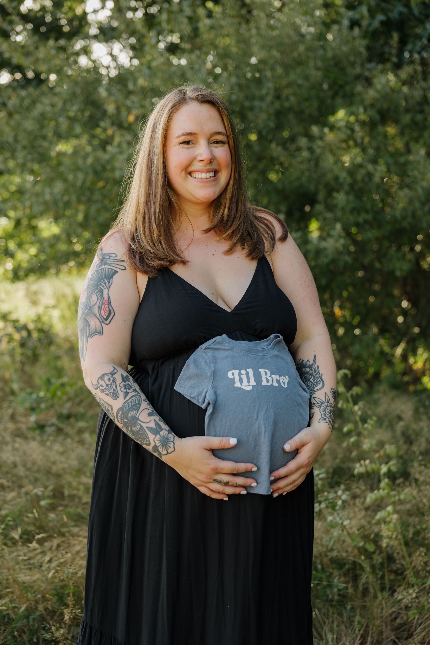 Smiling pregnant woman in black dress holding a blue Lil Bro onesie outdoors, showing tattoos on both arms.
