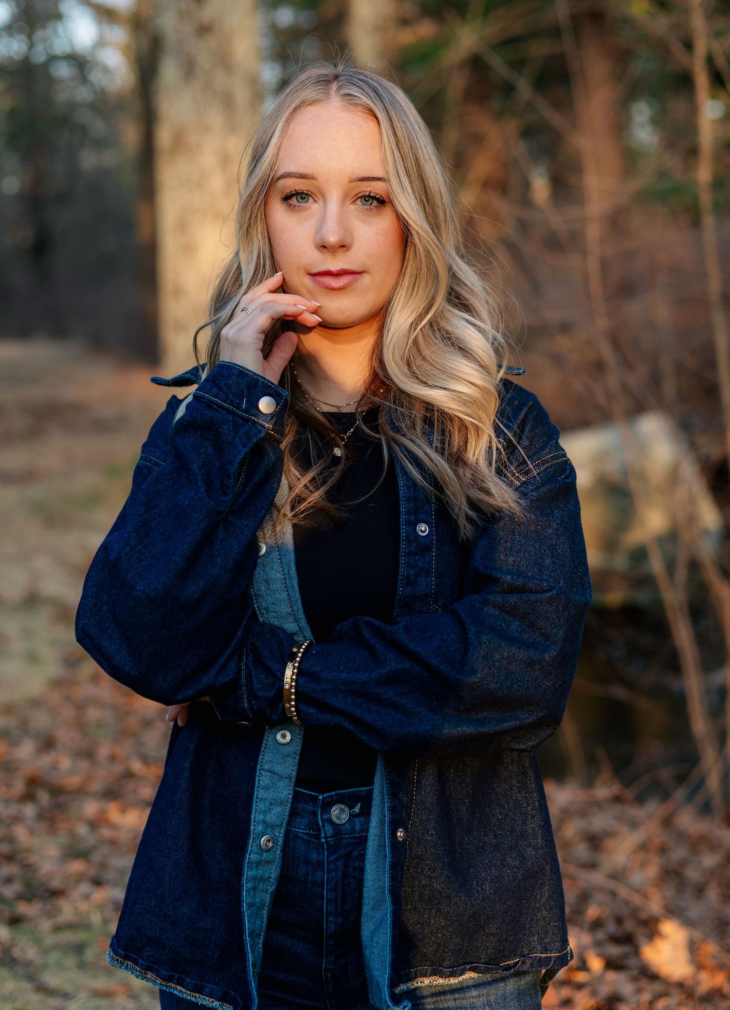 Blonde woman in dark denim jacket posing thoughtfully in sunlit wooded outdoor setting.