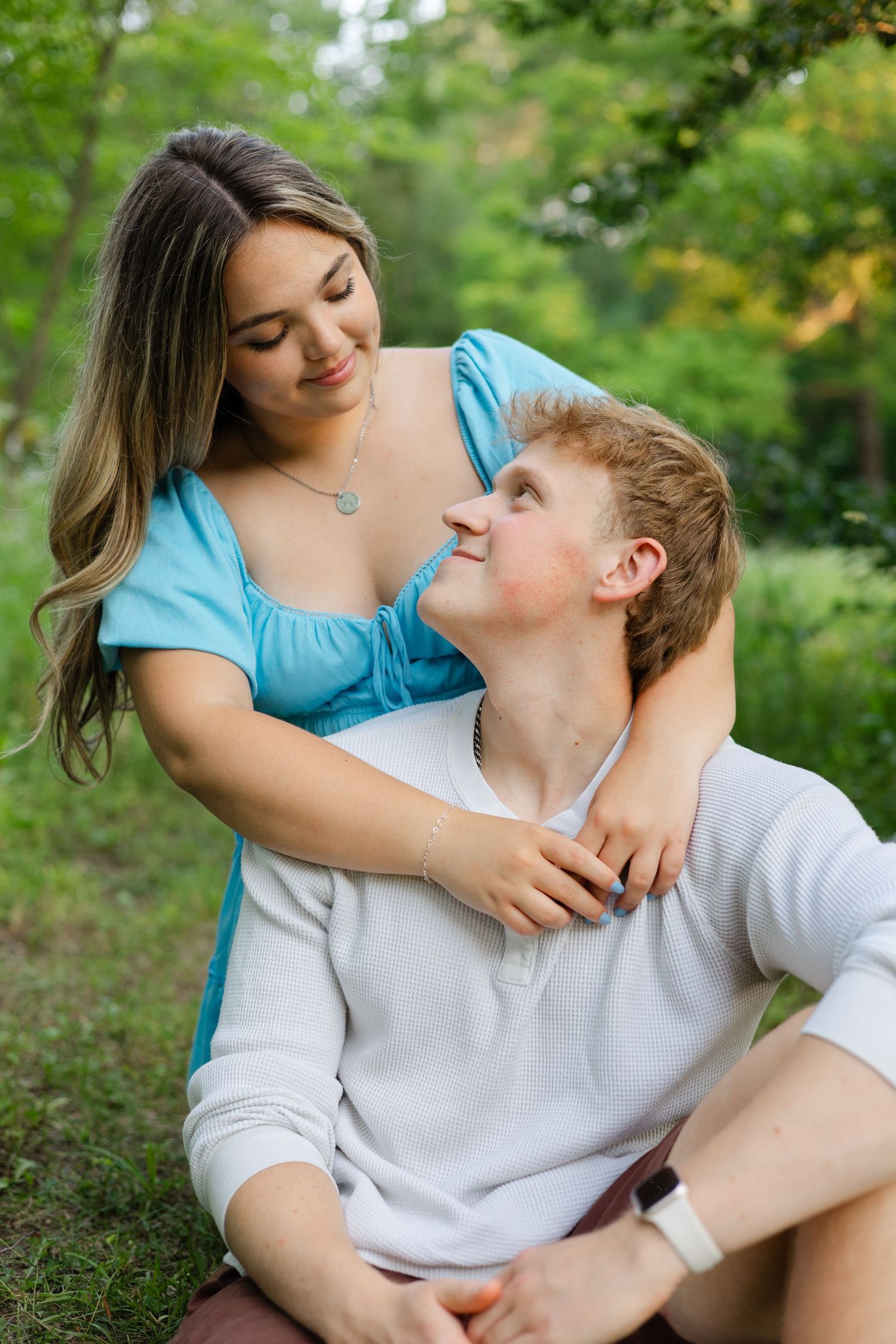A young couple shares a tender moment outdoors, with a woman in a blue dress embracing a man in white.