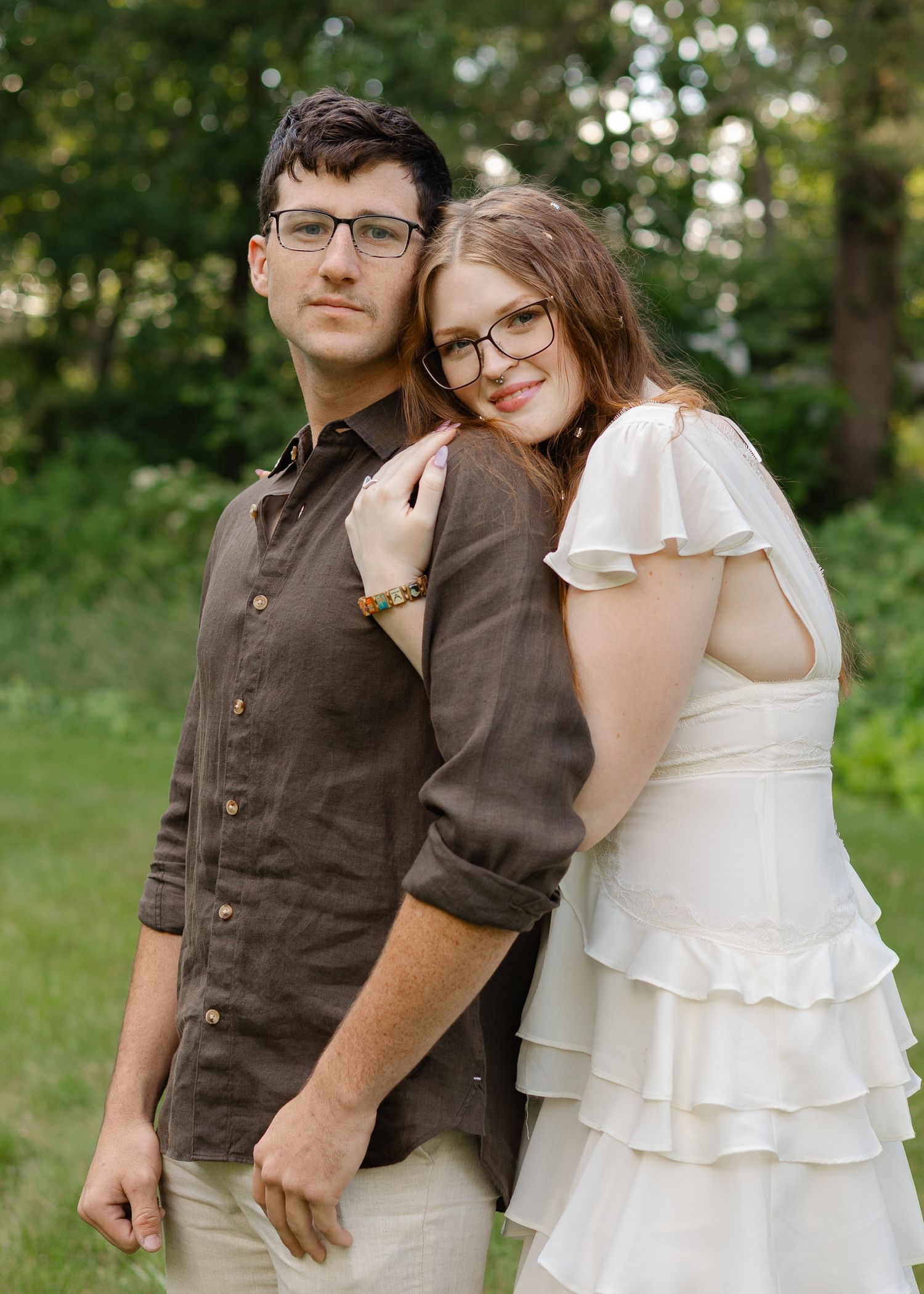 A couple poses together outdoors, man in brown linen shirt and woman in white ruffled dress, both wearing glasses.