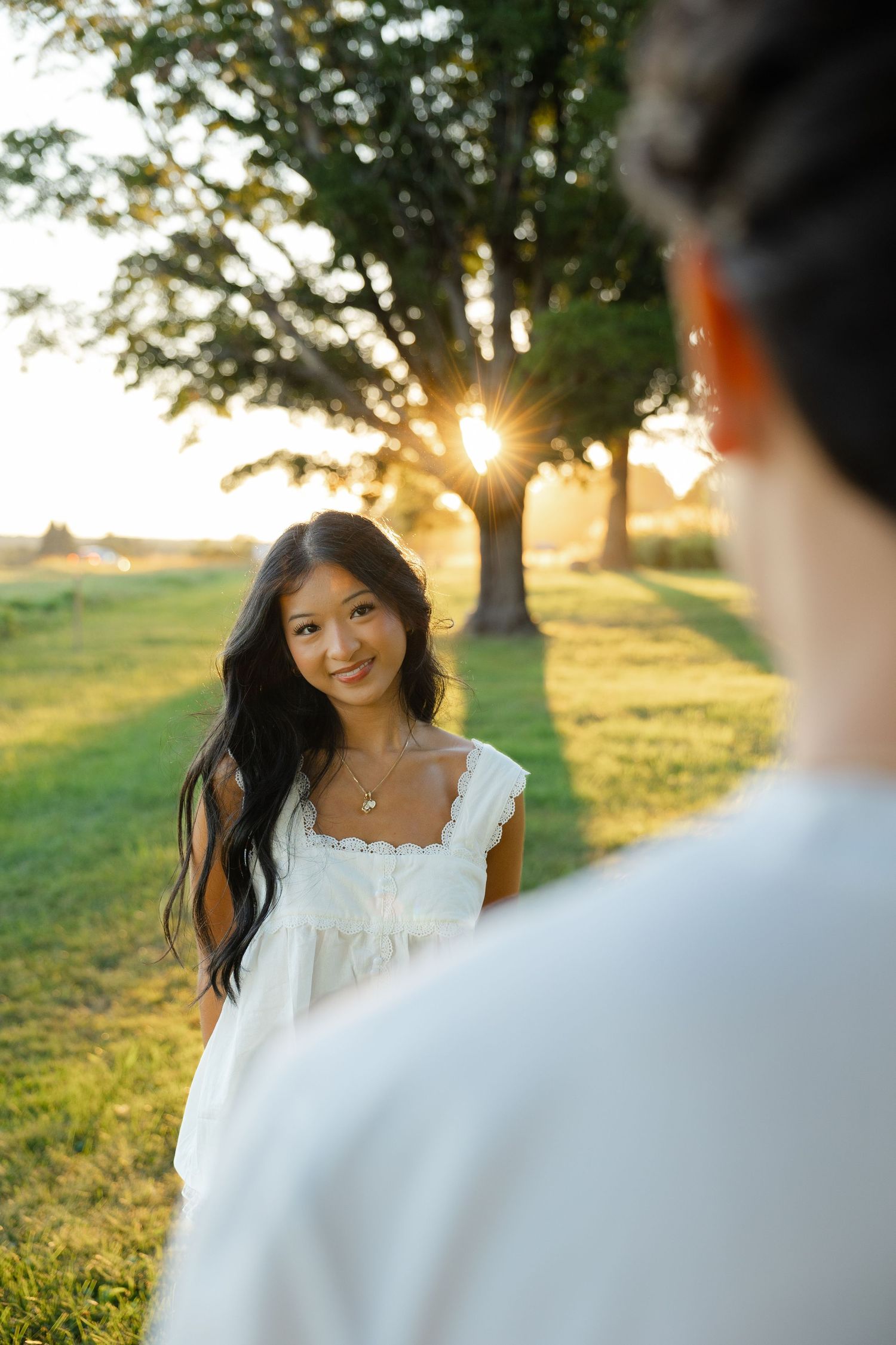 Young Asian woman in white dress smiling outdoors at golden hour with sunlight through trees.
