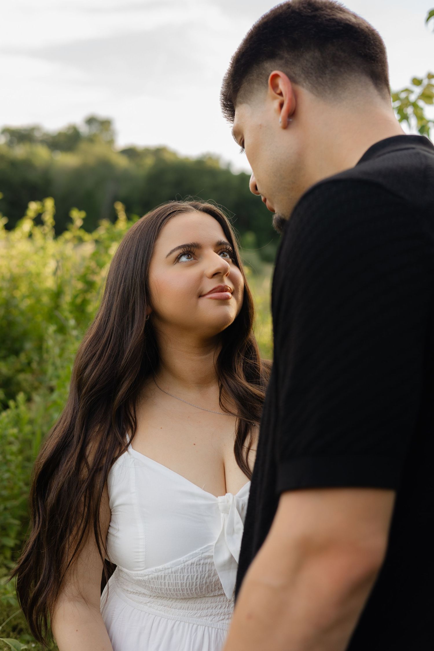 A young couple gazes at each other outdoors, woman in white dress with long dark hair, man in black t-shirt, green nature background.