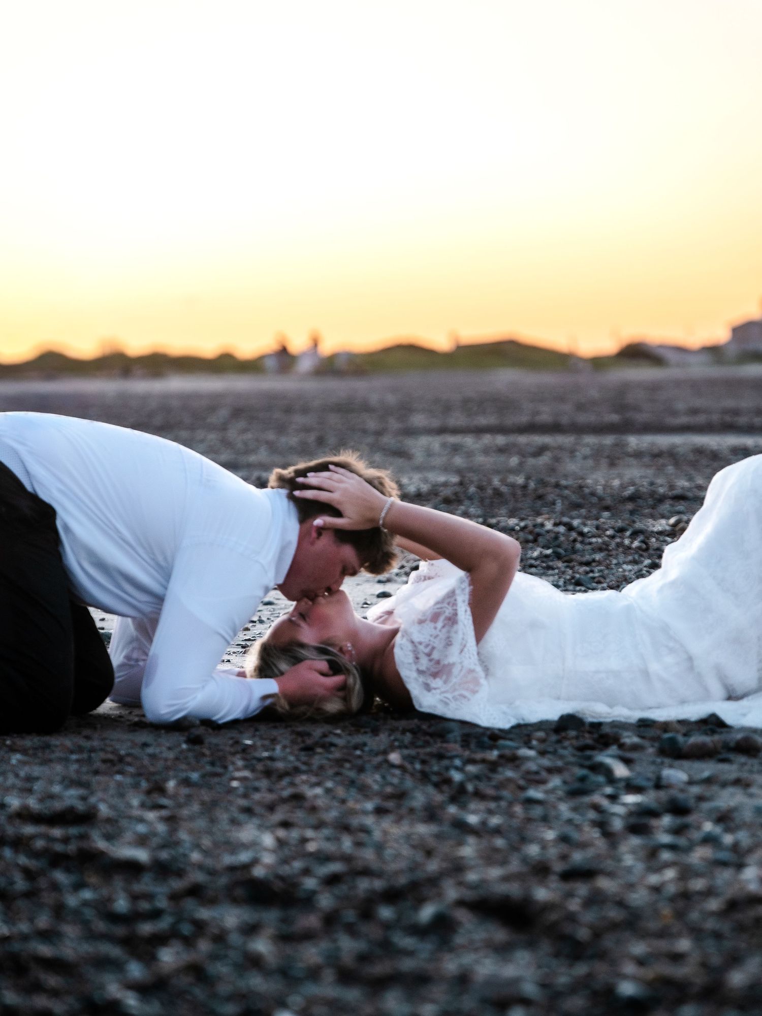 Couple in wedding attire lying on rocky beach sharing a kiss at golden sunset.