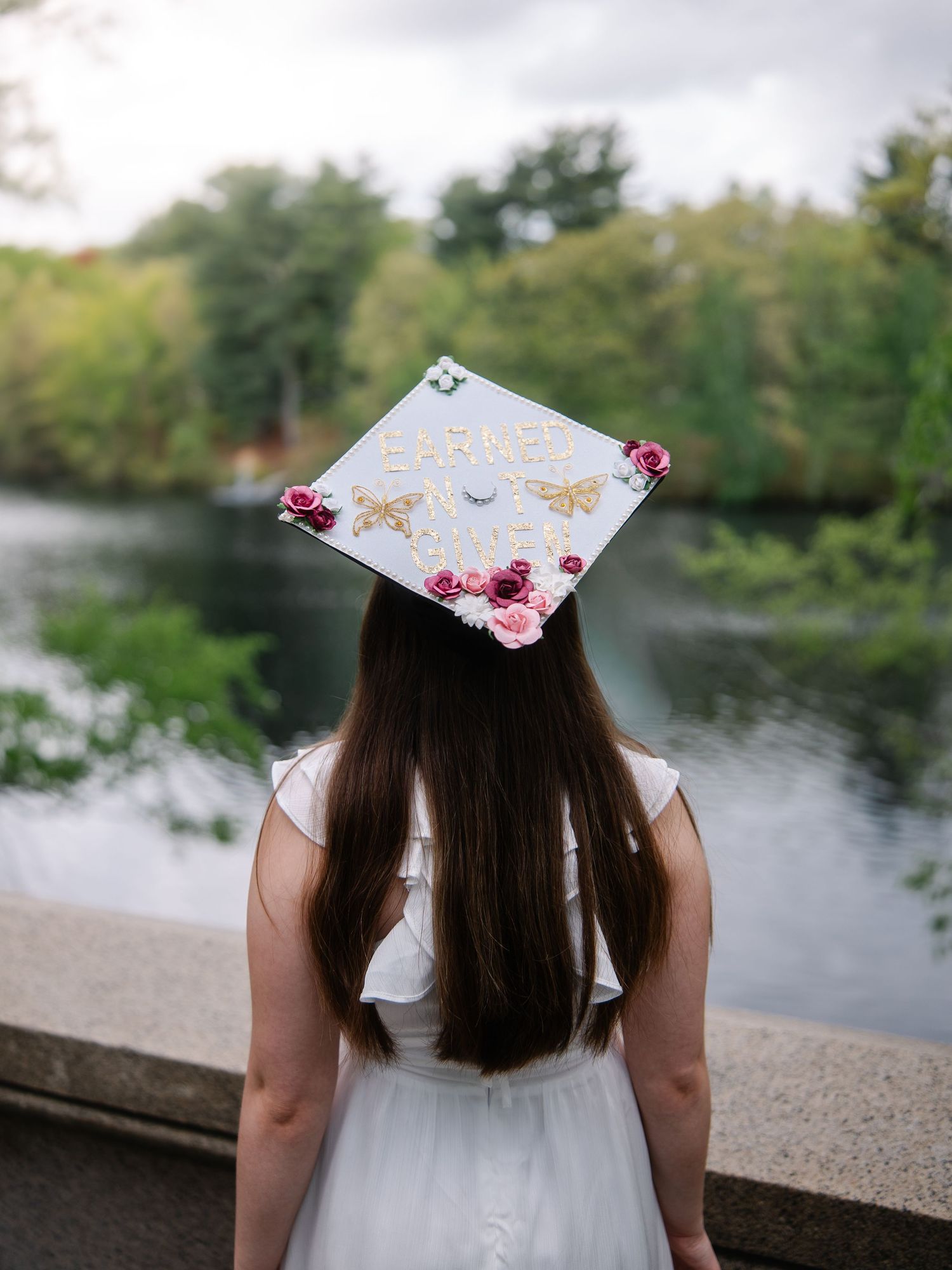 Graduate wearing decorated cap reading Earned Not Given with flowers, standing by a peaceful river.