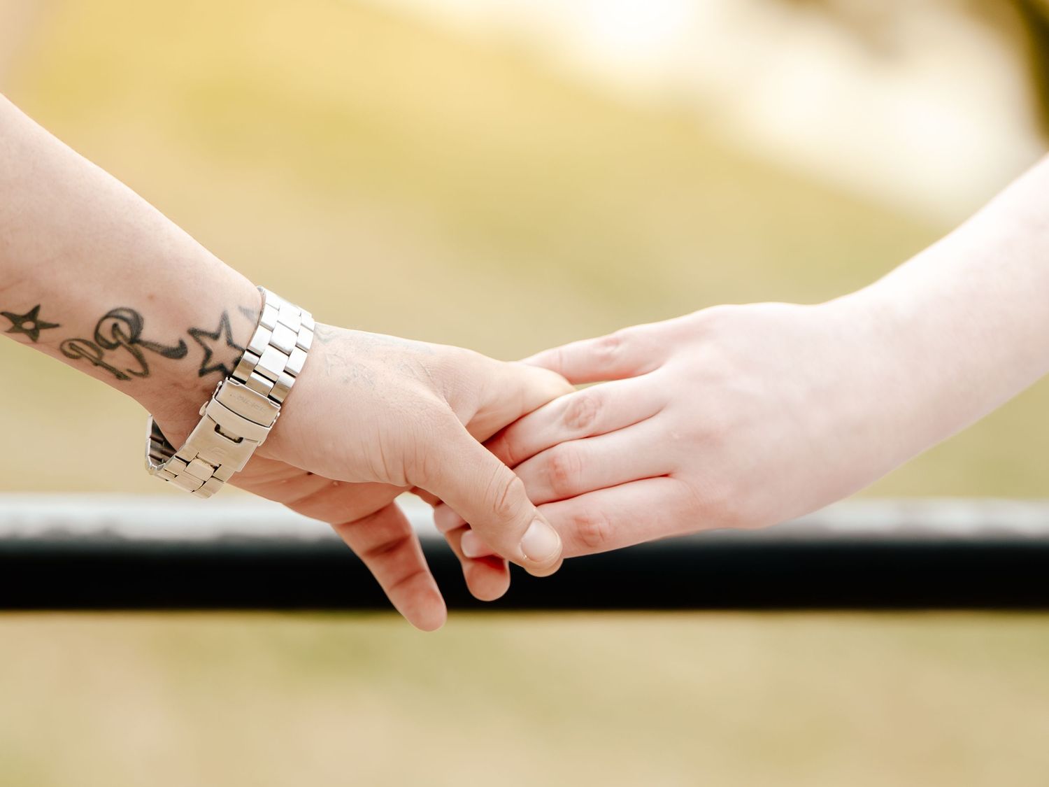 Two hands gently holding, one with a silver watch and star tattoos, against a soft bokeh outdoor background.